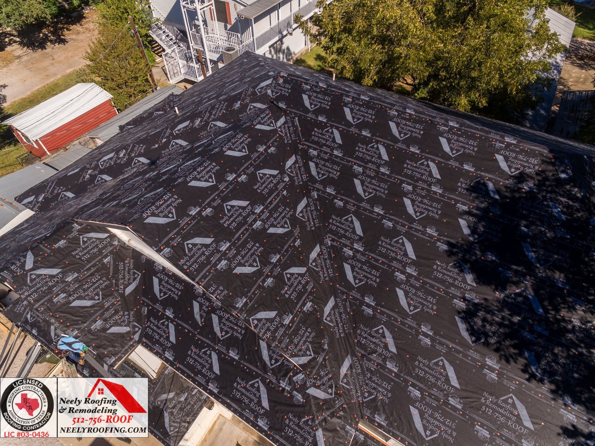 Aerial view of a black roof covered with multiple white repairs and patches near trees and houses