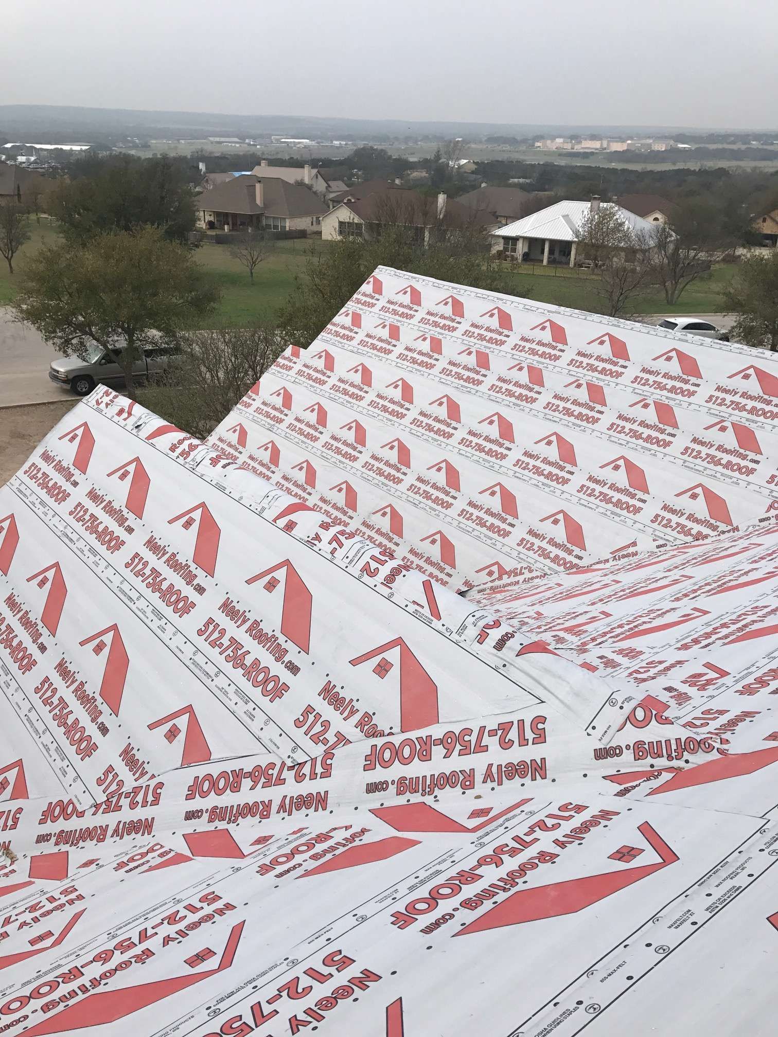 Roof covered in red-and-white protective wrap, overlooking suburban houses and a cloudy horizon.