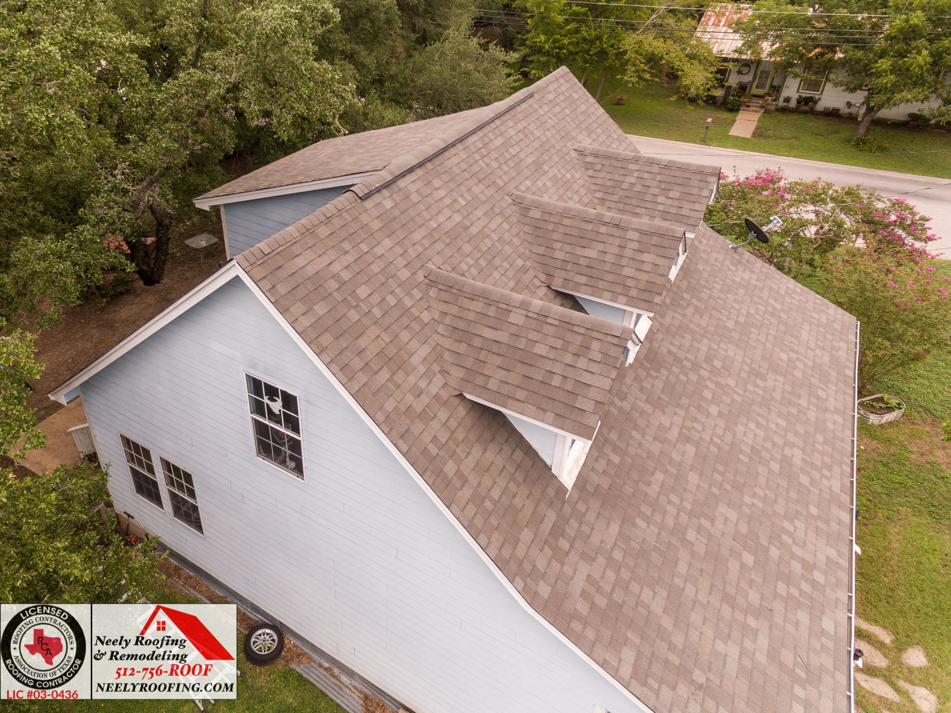Aerial view of a house with a gray shingle roof and white siding, surrounded by trees and grass.