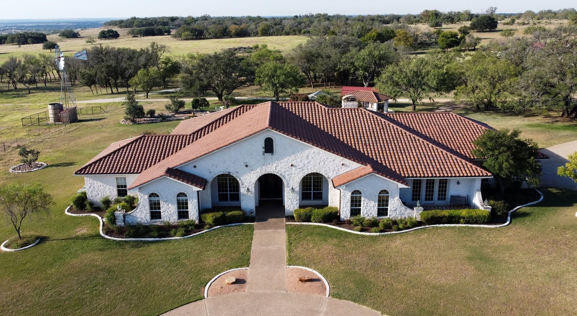 Aerial view of a white stucco house with red tile roof, arched entry, and landscaped lawn in a rural setting