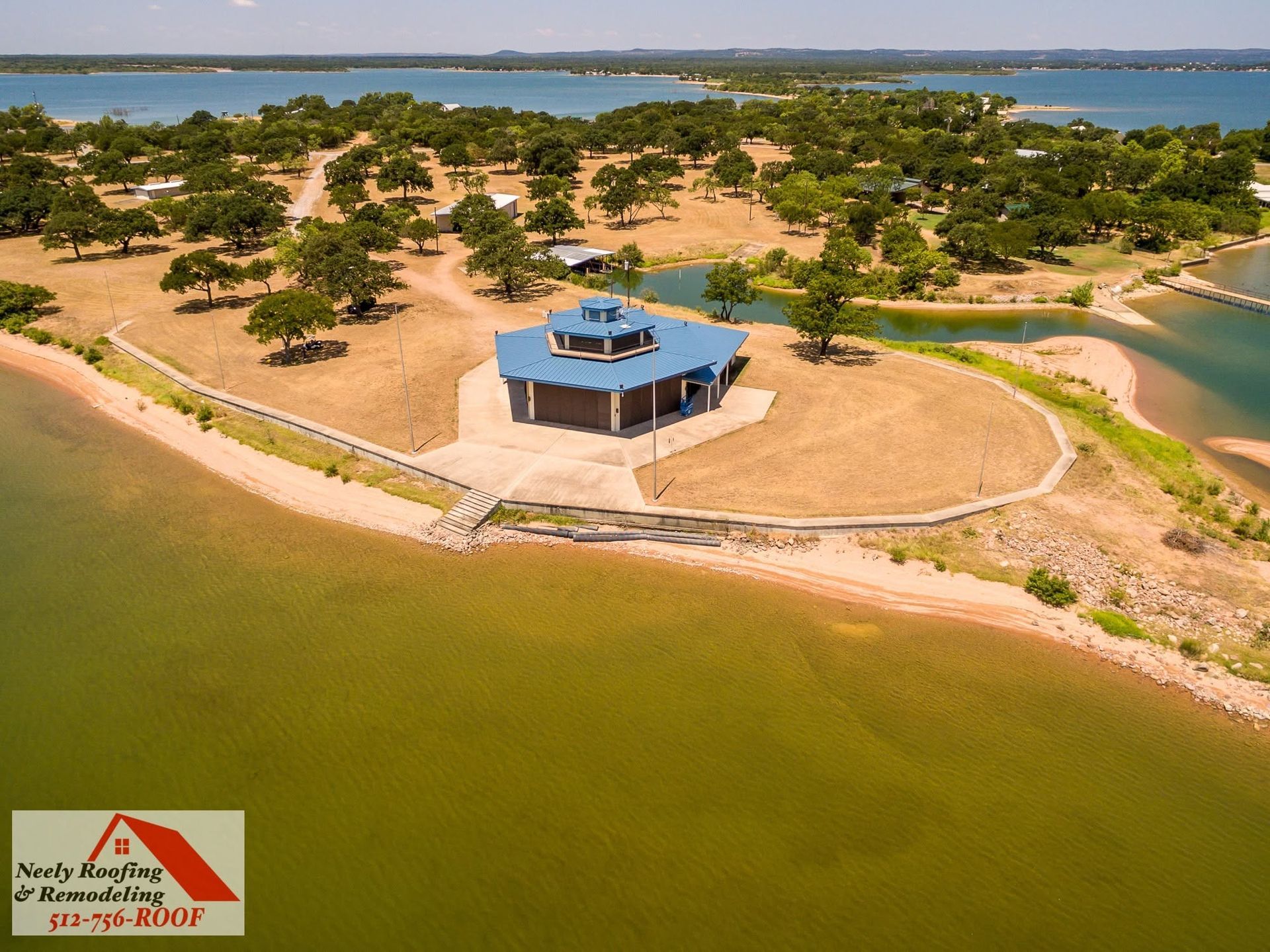 Aerial view of a blue-roofed gazebo on a sandy peninsula beside green water and trees