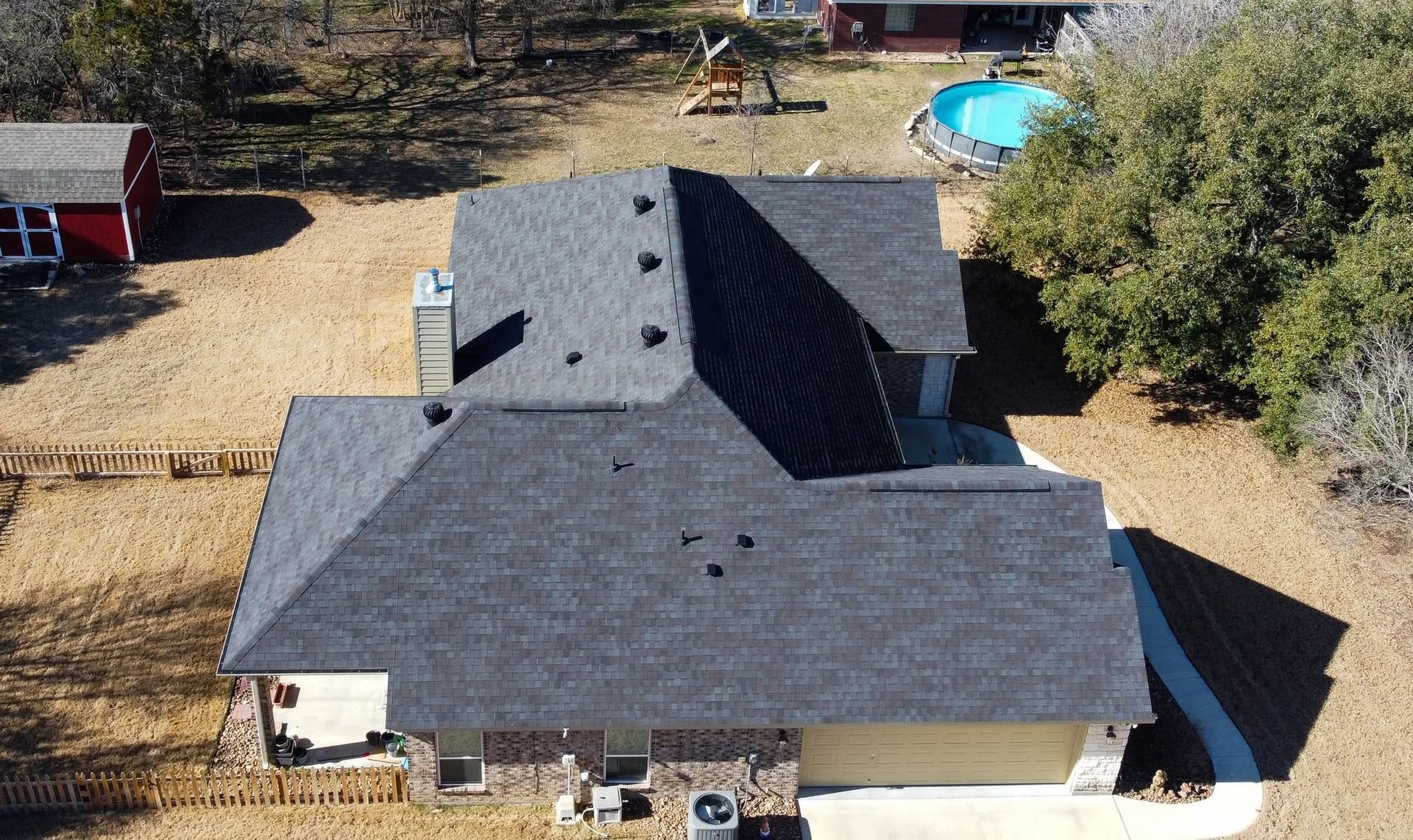 Aerial view of a house with a dark gray roof and surrounding yard, with a small pool in the background.