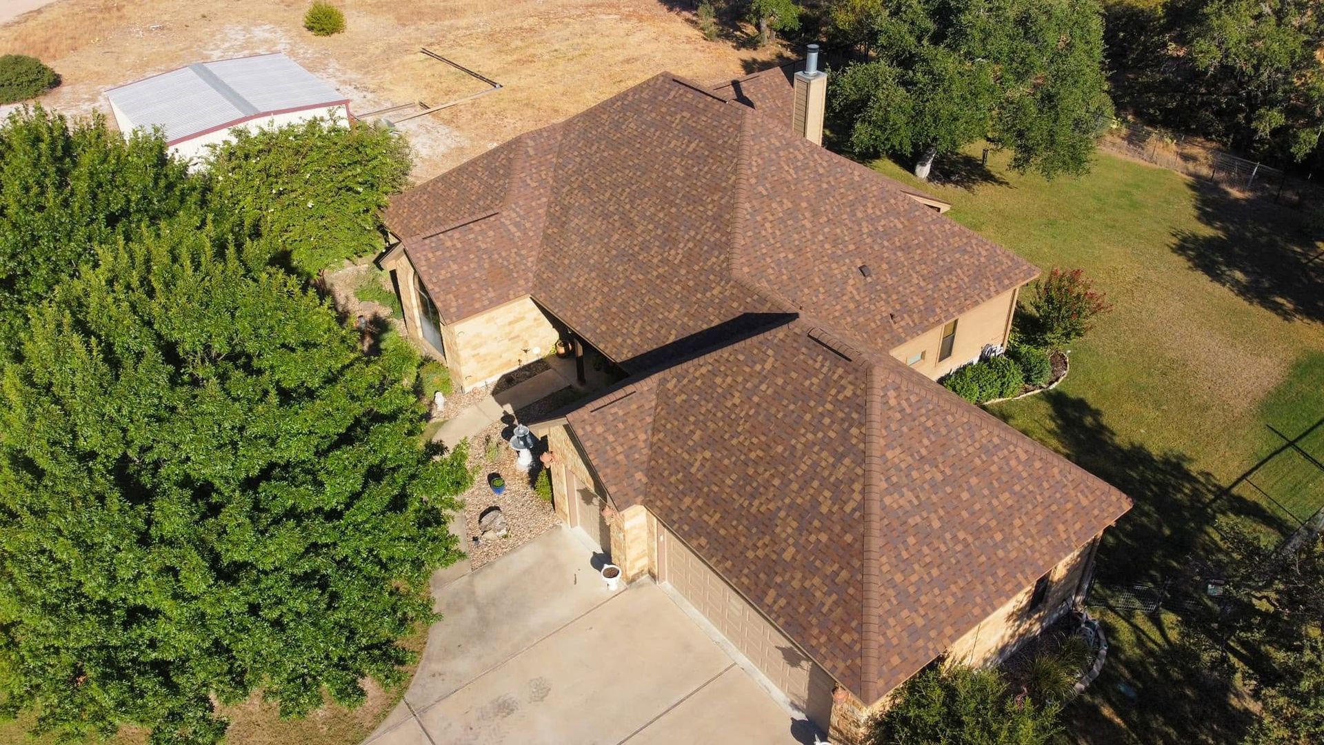Aerial view of a brown-roofed house with a driveway, surrounded by trees and yard.