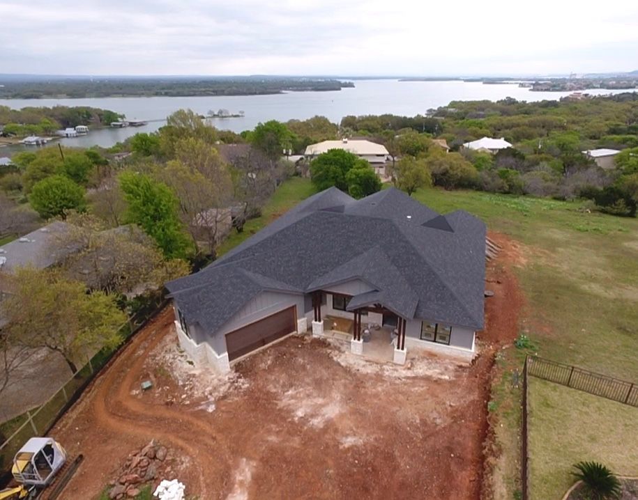Aerial view of a gray-roofed house with a garage, dirt yard, and water in the background.