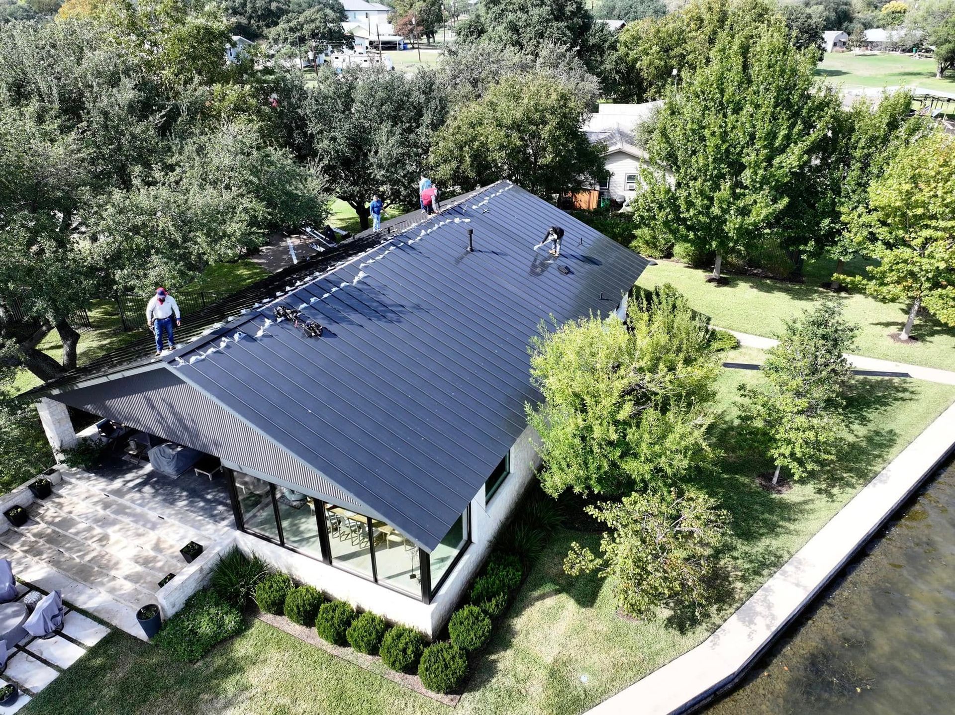 Aerial view of a house with a dark roof, surrounded by trees and a lawn, with a few people on the roof.