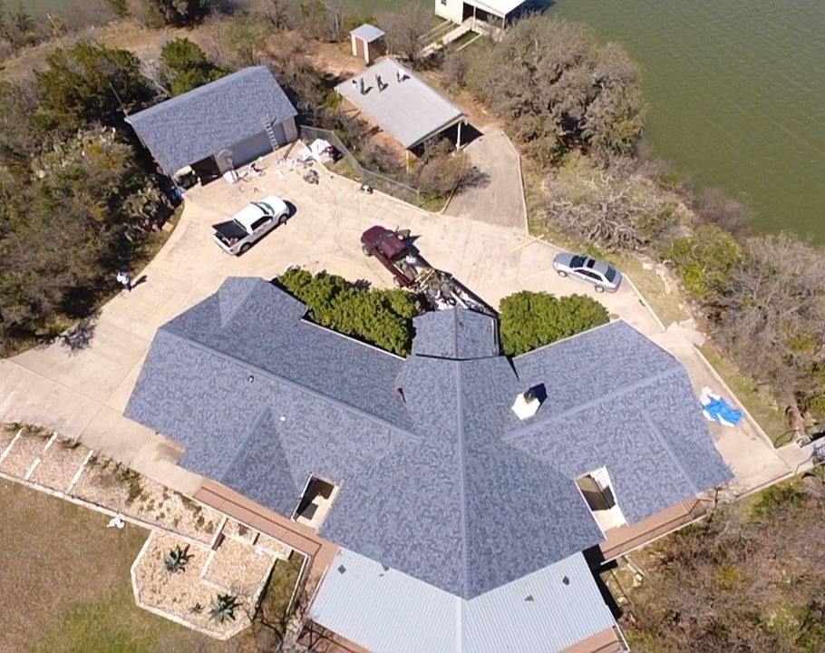 Aerial view of a large gray-roofed house with parked vehicles and surrounding trees and dirt driveway