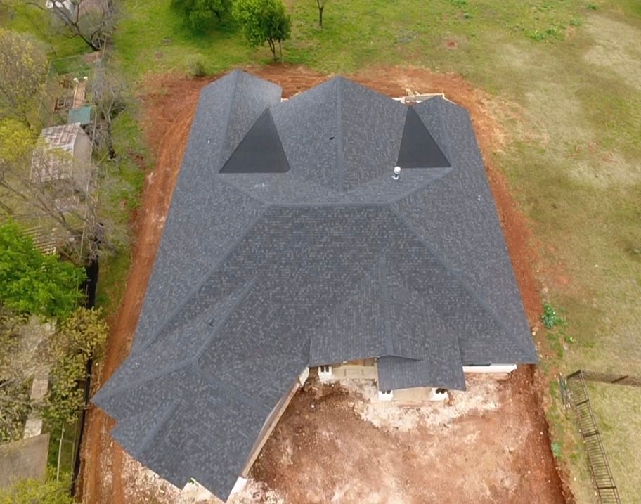 Aerial view of a house with a dark gray roof and unfinished front yard surrounded by grass and trees