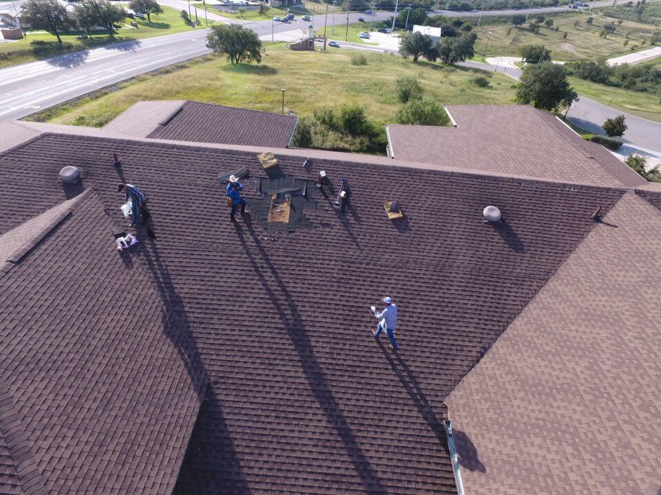 Workers on a brown shingled roof near a road, using tools during roof maintenance.