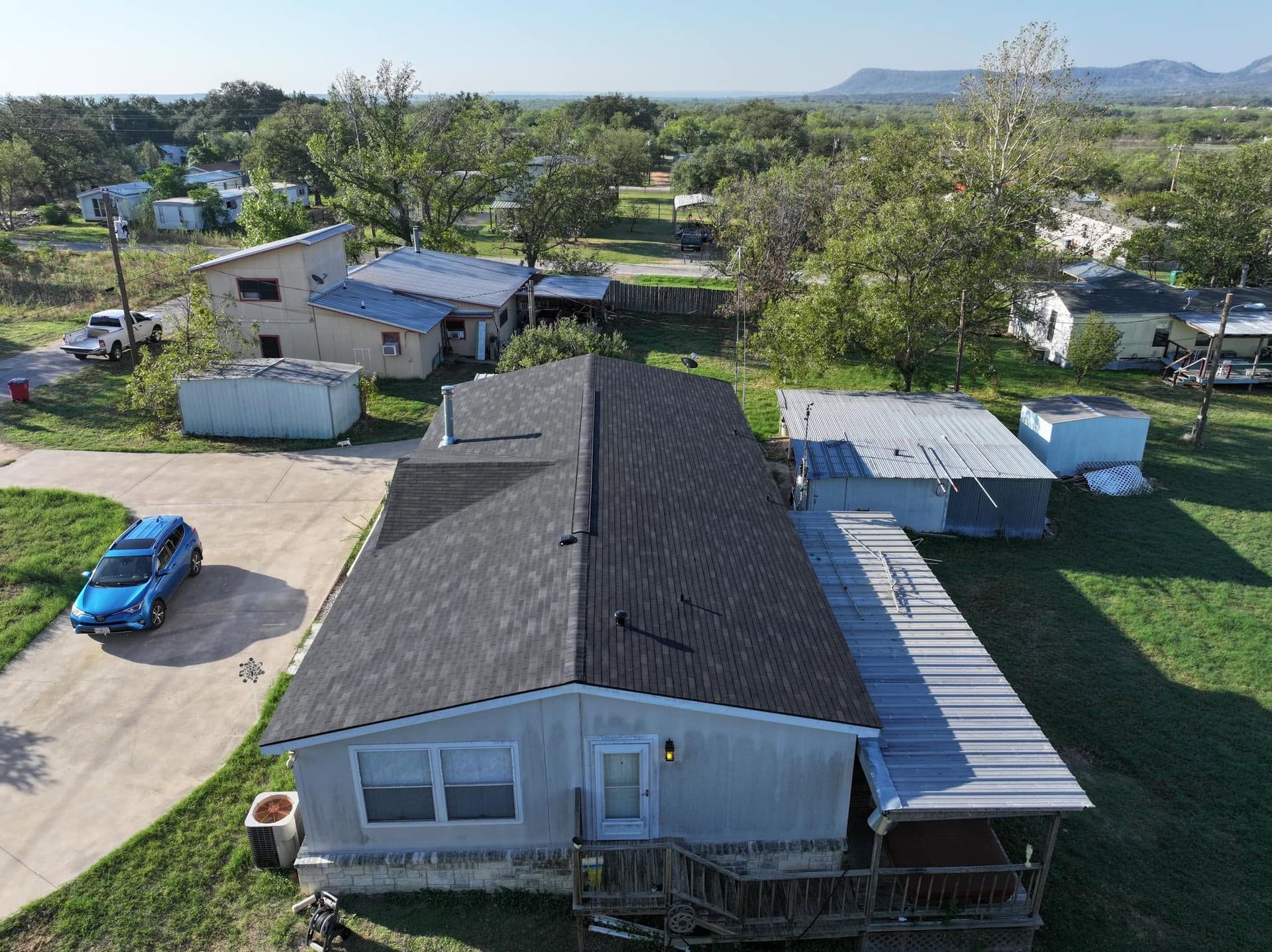 Aerial view of a suburban house with a dark roof, blue car, and green yard with trees and mountains beyond.
