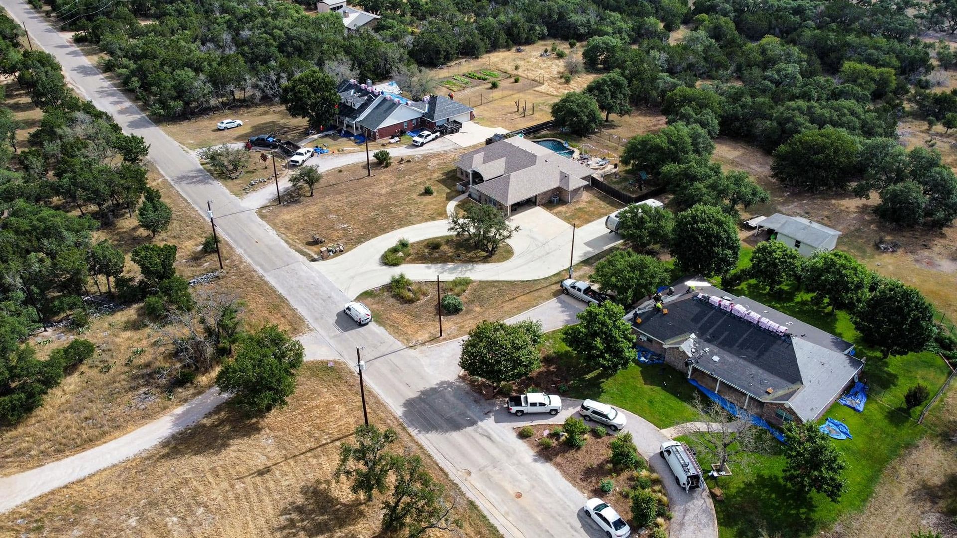Aerial view of a suburban road with several houses, driveways, parked cars, and trees in a wooded area