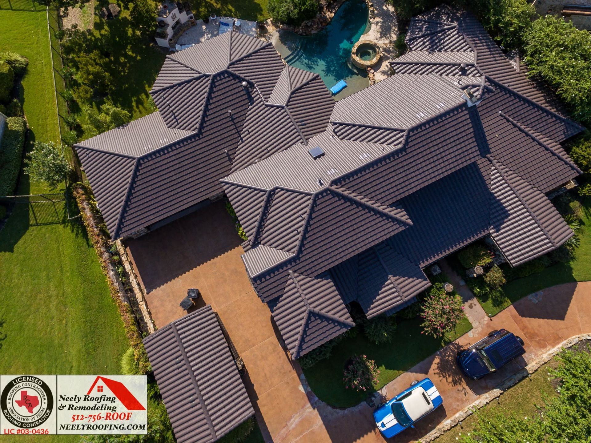 Aerial view of a large dark-roofed house with a driveway, pool, and parked cars surrounded by trees.