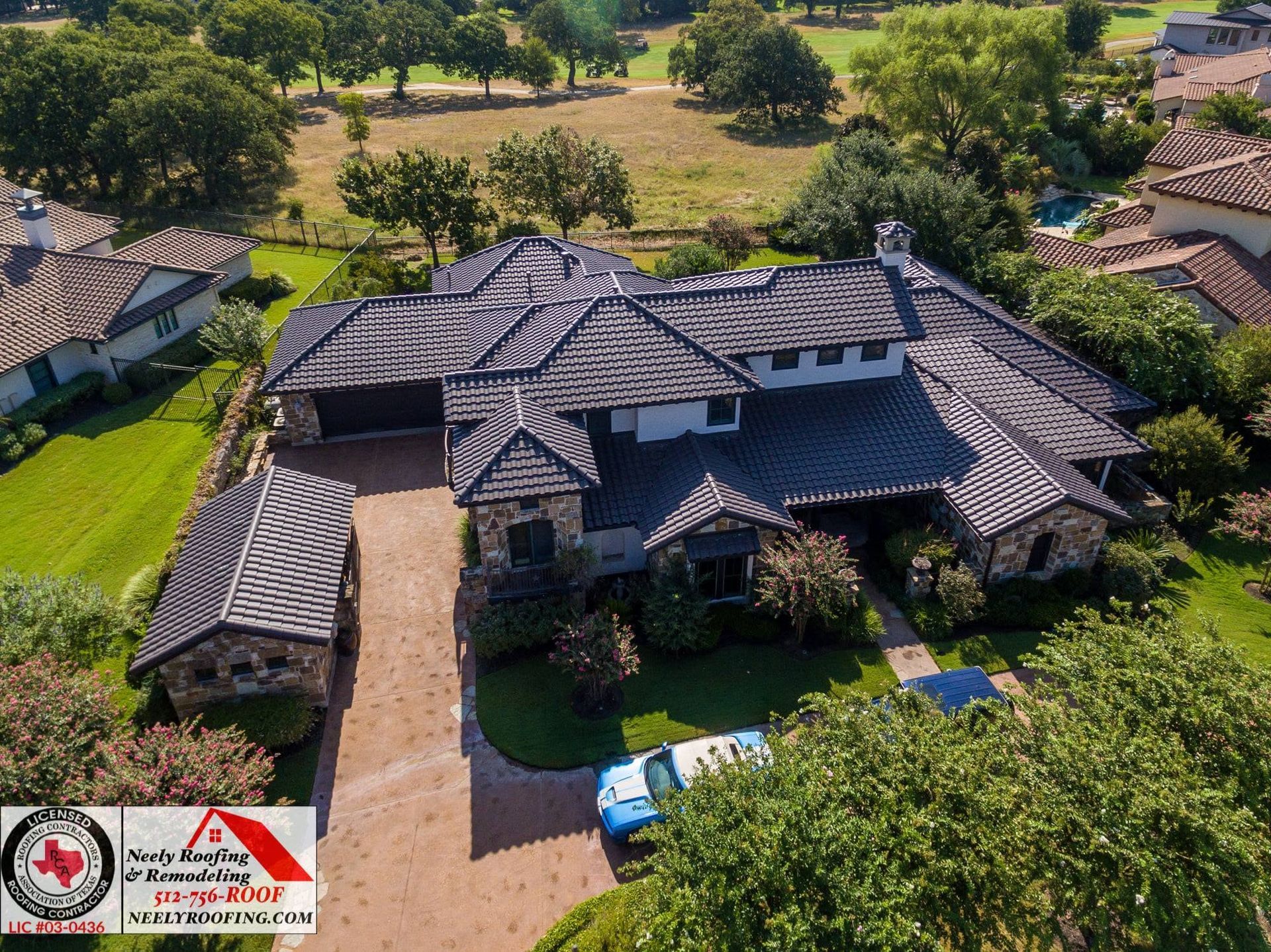 Aerial view of a large dark-roofed house with a driveway and trees surrounding it.