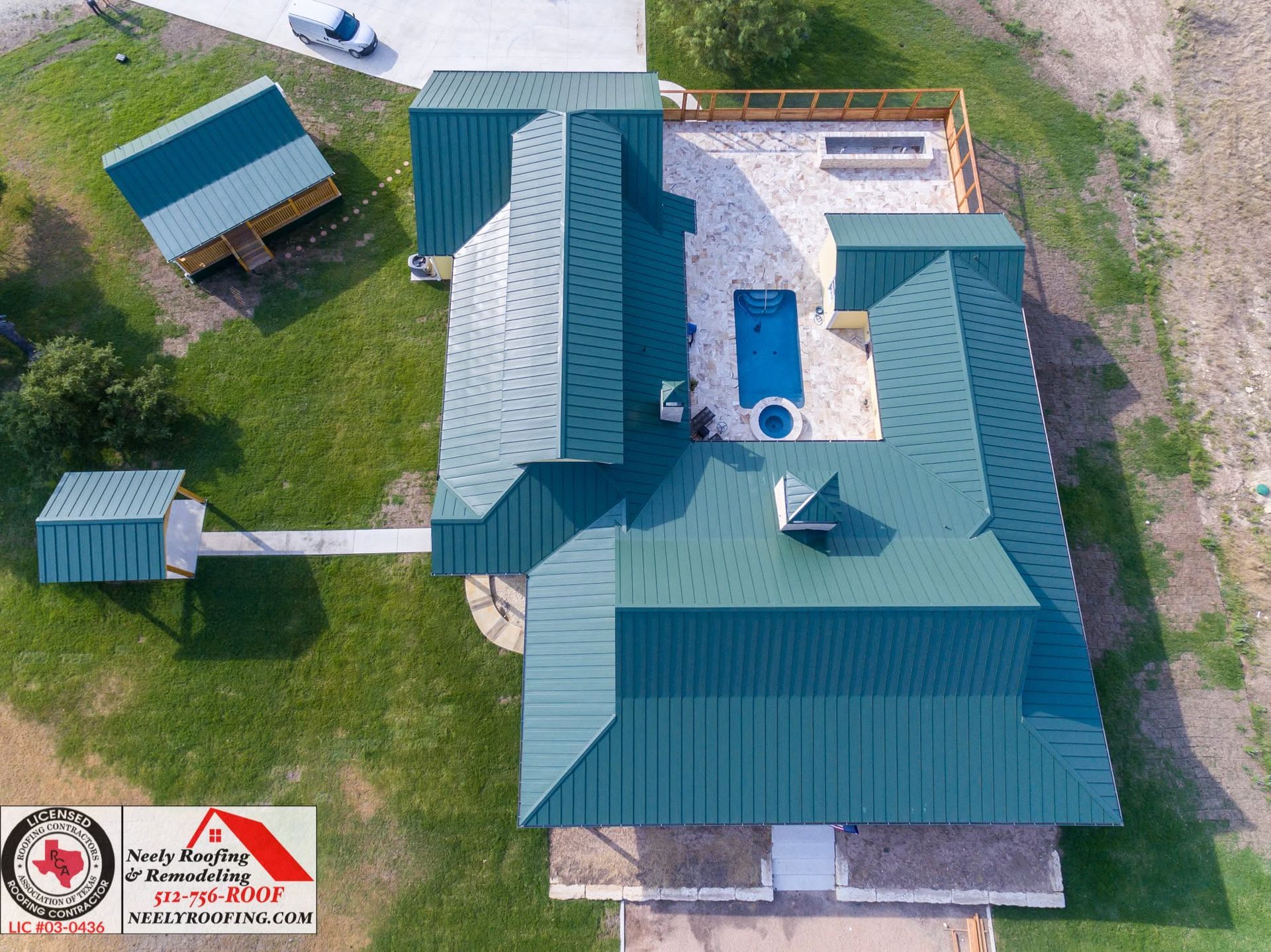Aerial view of a house with teal metal roofs, a central white courtyard, and a small outbuilding on a grassy lot