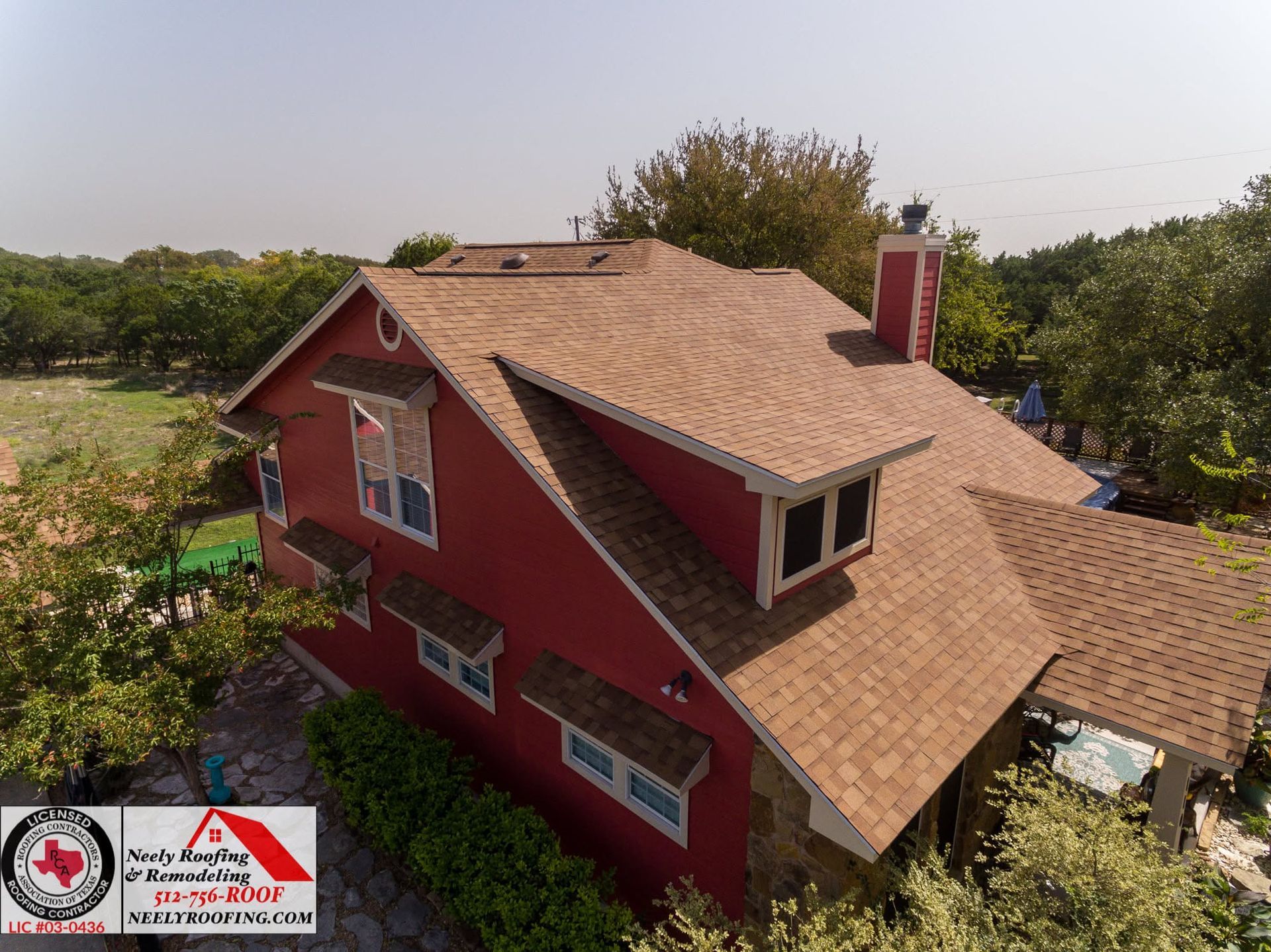 Aerial view of a red house with a tan shingle roof surrounded by trees and greenery.