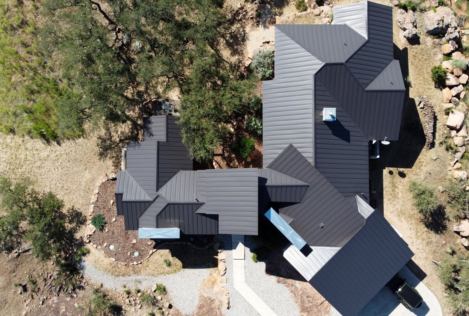 Aerial view of a dark gray-roofed house surrounded by trees and a rocky yard.