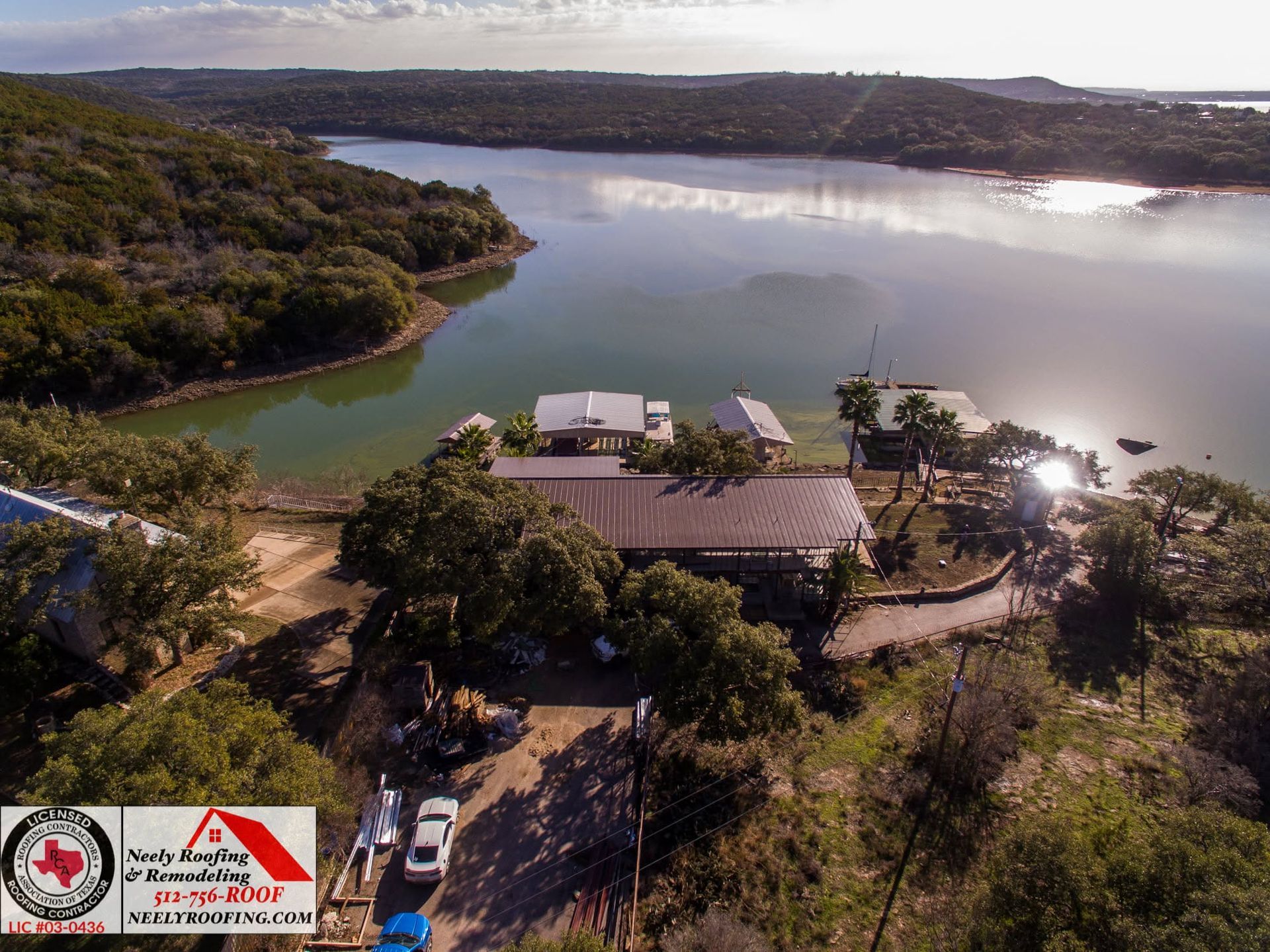 Aerial view of a lakeside building complex surrounded by trees with calm water and distant hills