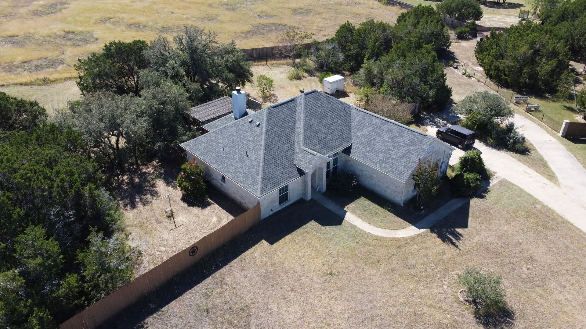 Aerial view of a gray-roofed house with a fenced yard, trees, and a curved driveway.