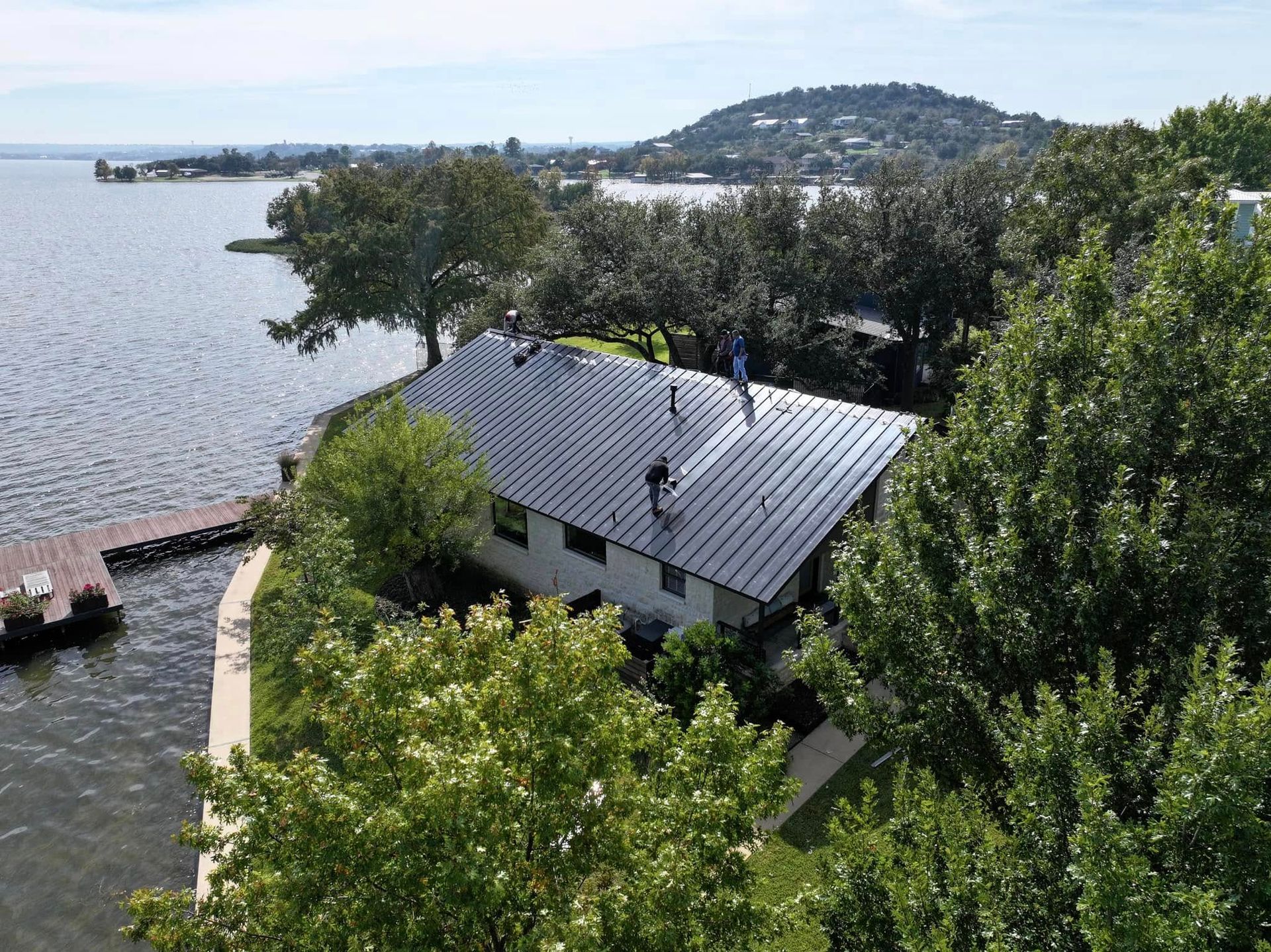 Shoreline house with gray roof beside calm water, surrounded by green trees and a distant hill