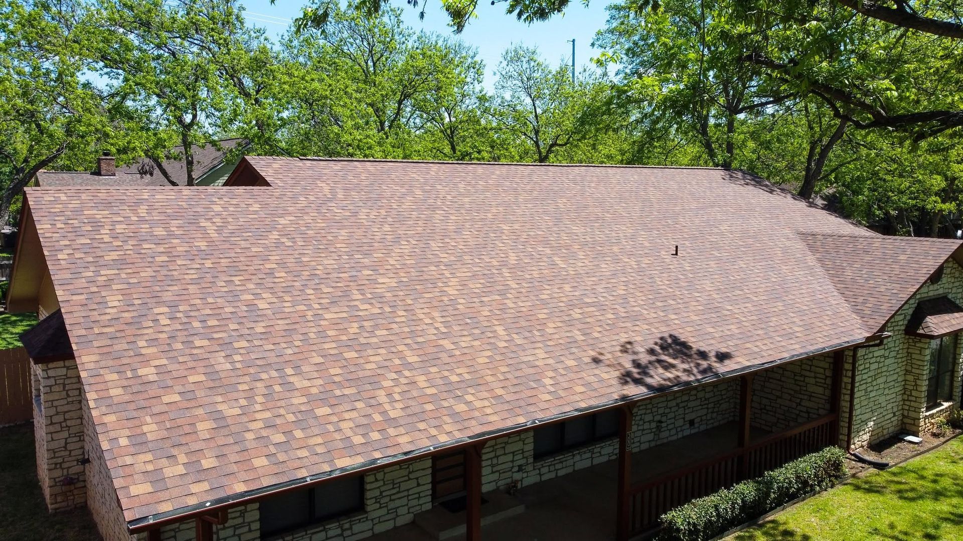 Aerial view of a large brown shingled roof surrounded by green trees and foliage.