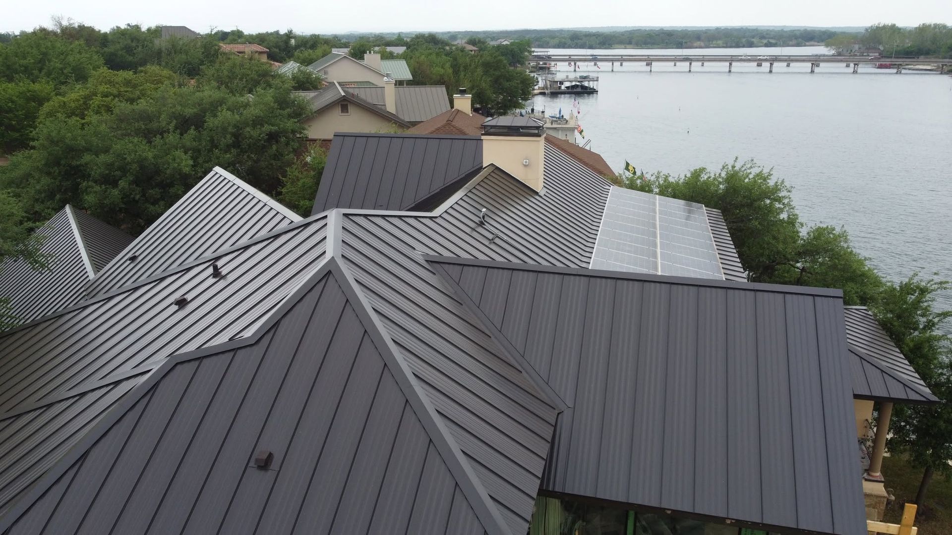 Metal rooftops beside a calm waterfront, with trees and a bridge in the distance.