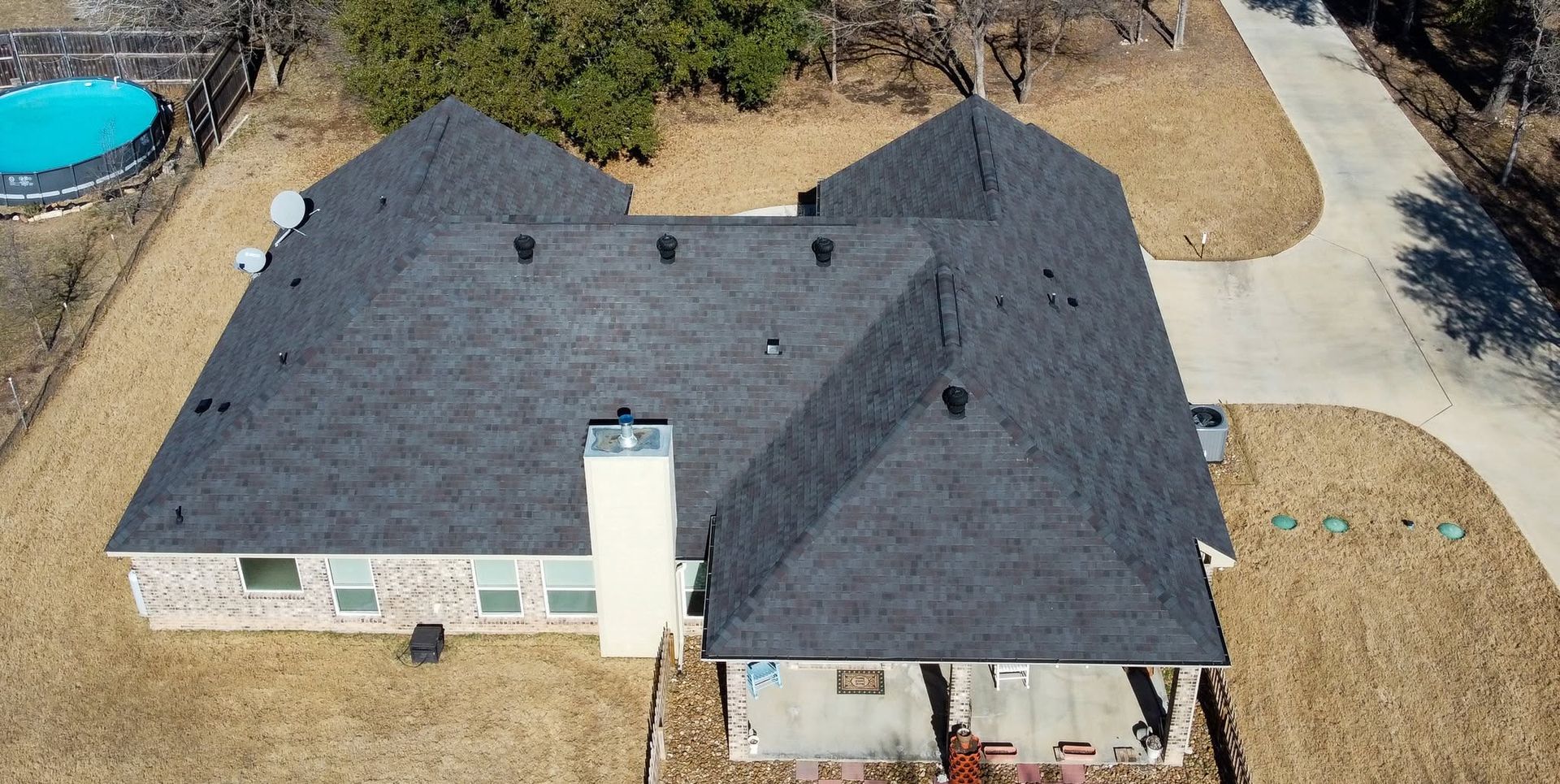 Aerial view of a gray shingle roof on a beige house with a chimney and yard.