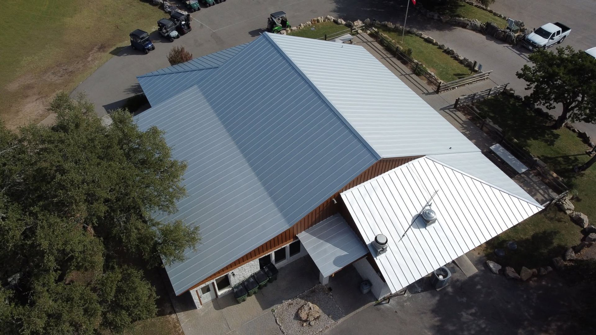Aerial view of a large metal-roofed building with white and gray roofs, trees, and parked cars nearby