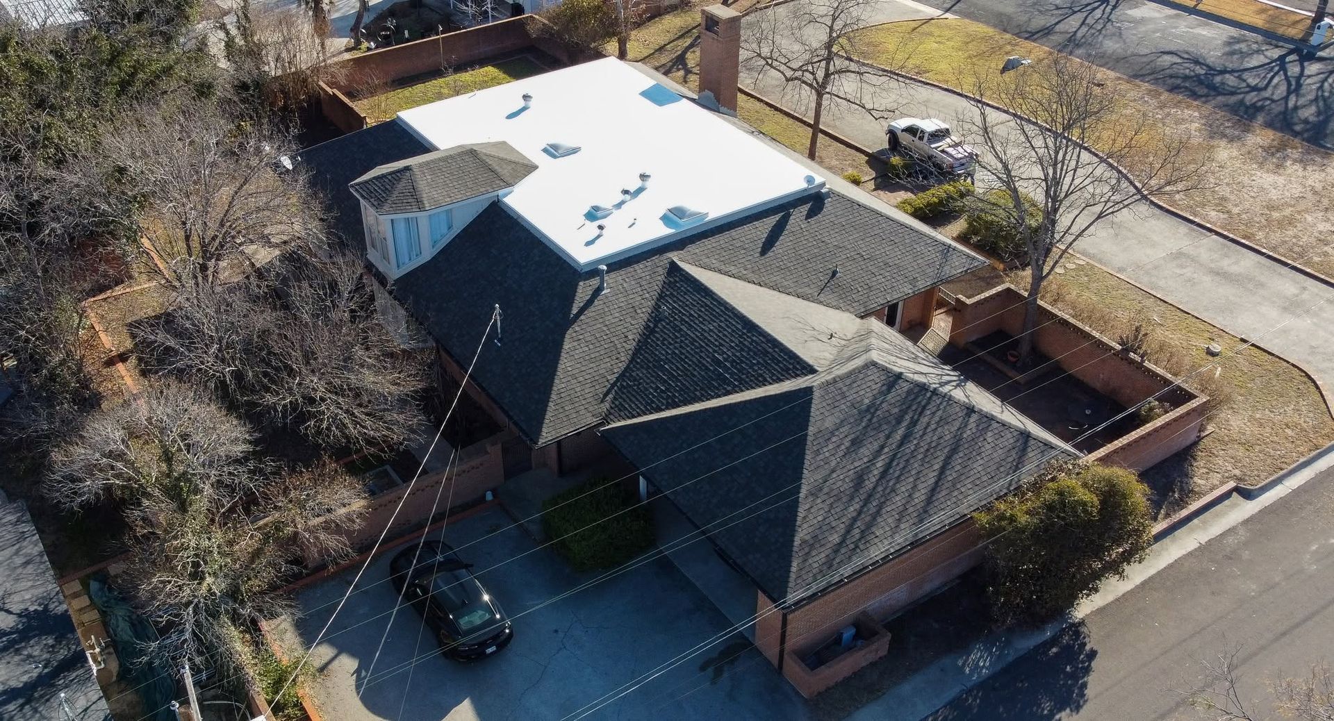 Aerial view of a dark-roofed house with a white rooftop, driveway, and parked cars beside a road.