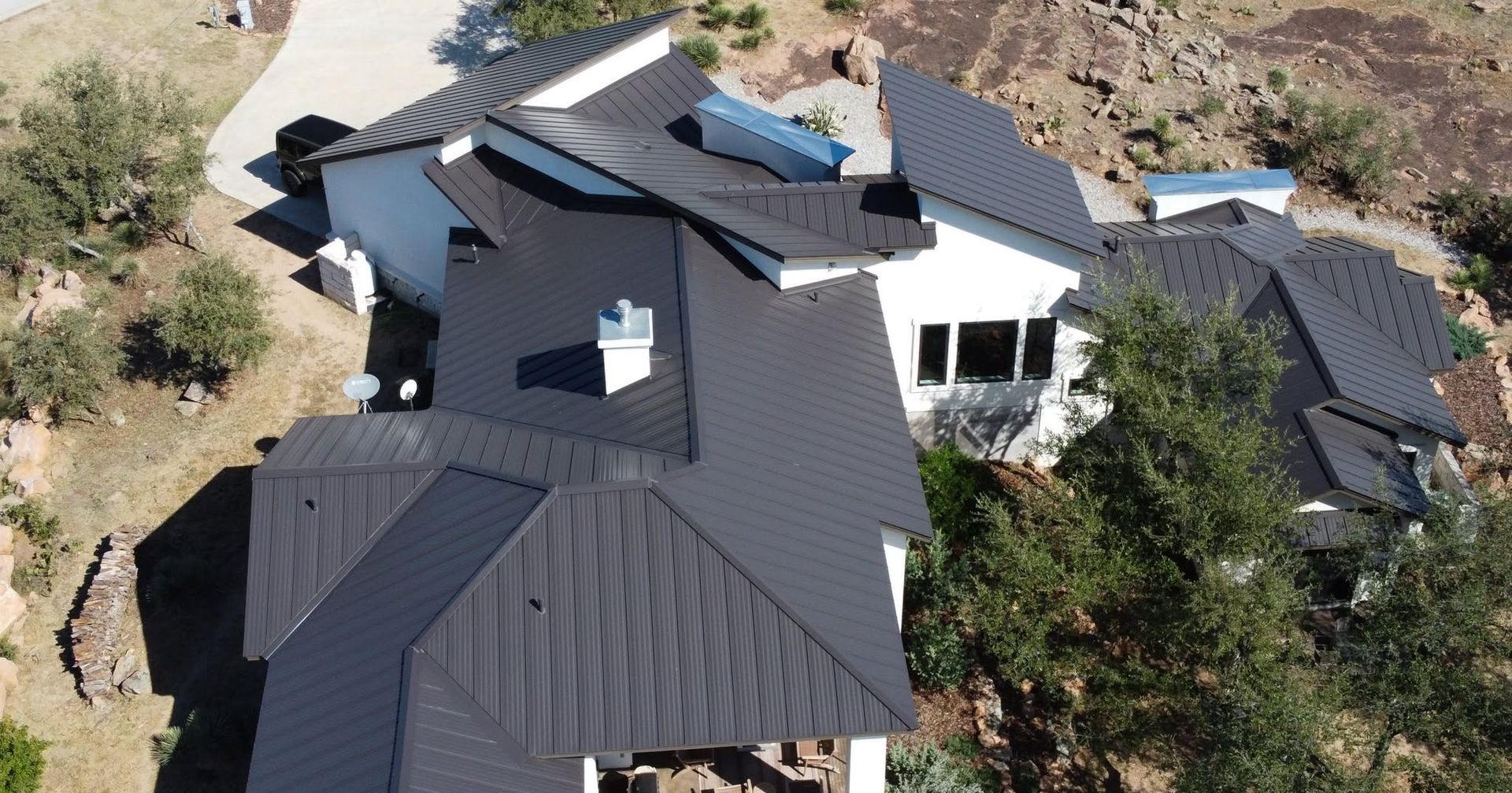 Aerial view of a dark-roofed house with multiple gables surrounded by trees and rocky terrain.