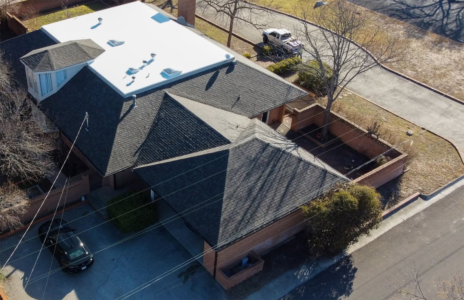 Aerial view of a brick building with dark roofs, a white rooftop, and a parked car beside the street.