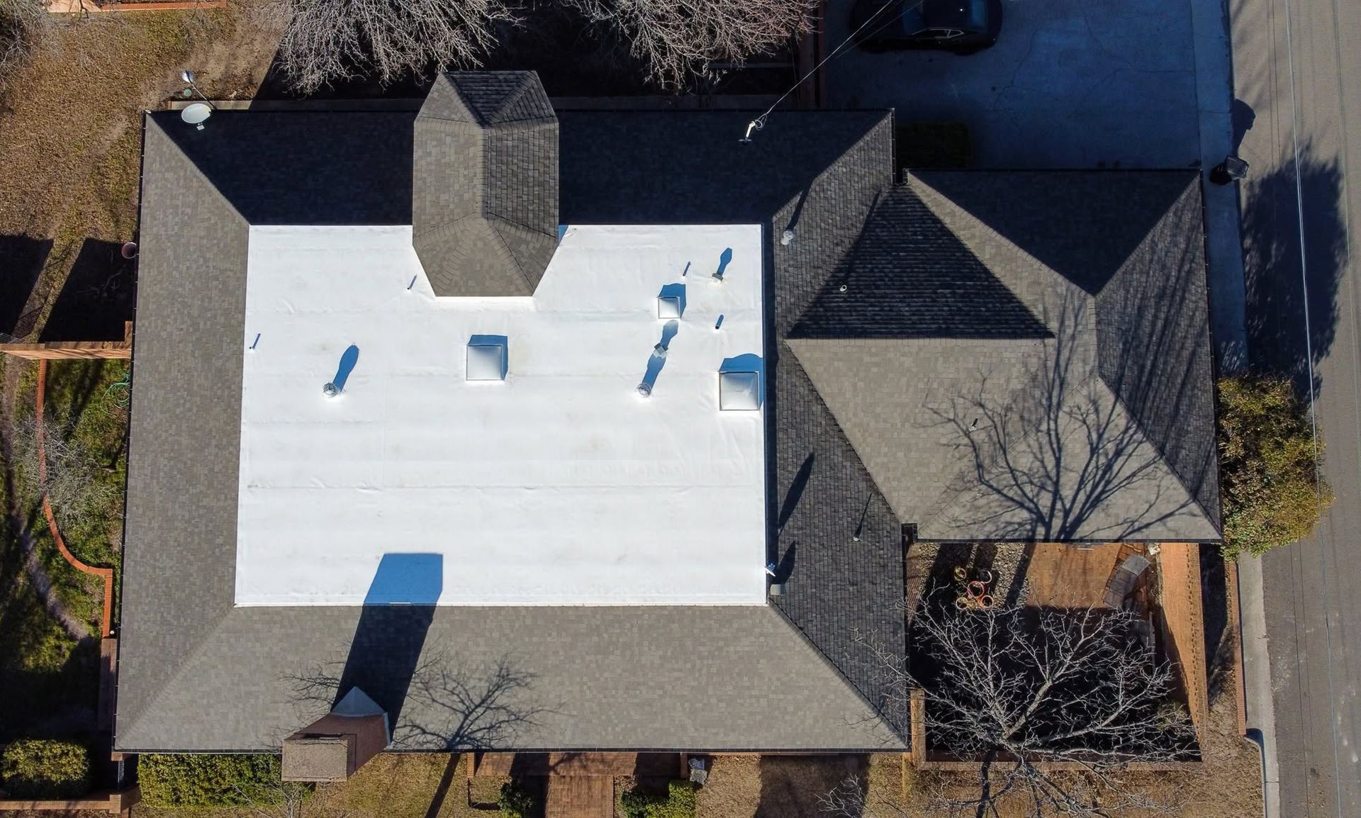 Aerial view of a house with a white roof, dark gray sections, and surrounding trees.