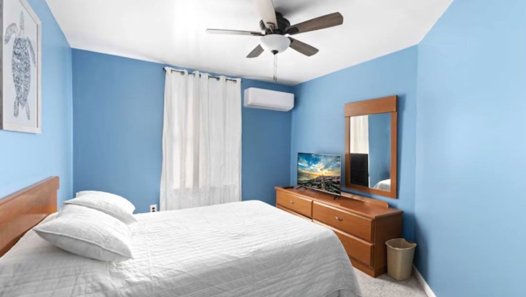 A bedroom with blue walls, a bed, a dresser, a television, and a ceiling fan.