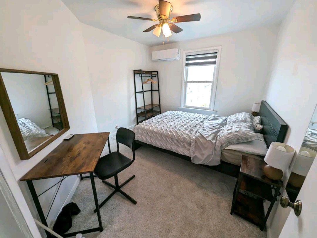 A bedroom with a bed, desk, chair, mirror, and ceiling fan.