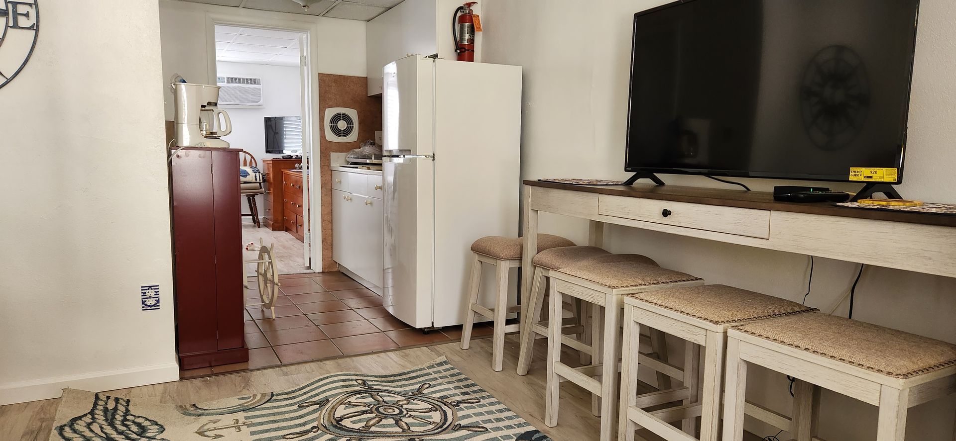A small apartment interior with a kitchen, living area with a TV, stools, and a rug.
