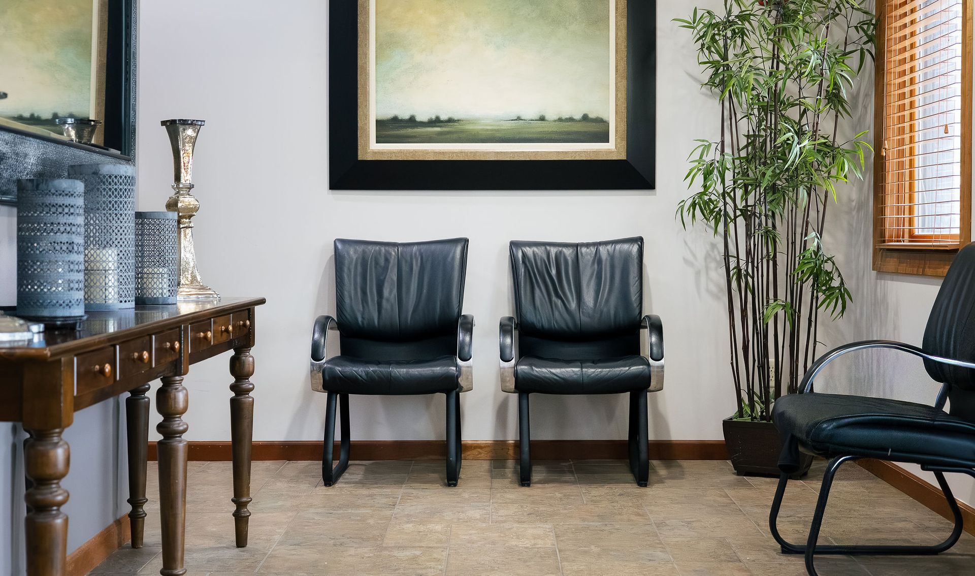 Waiting area with two black leather chairs beneath a painting. Console table with decor on left, bamboo plant on right.
