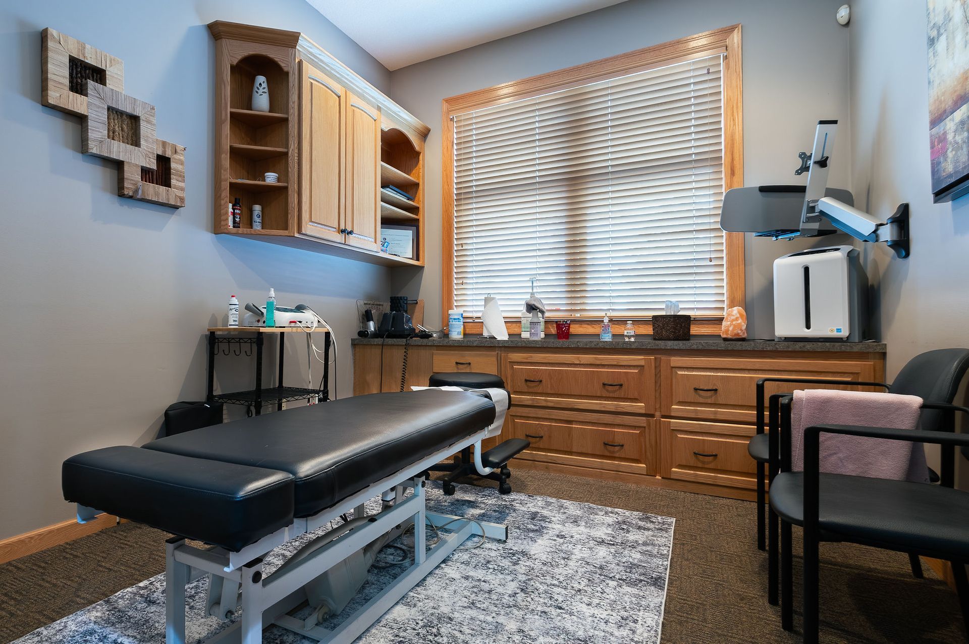 Chiropractor's office: treatment table, cabinetry, window with blinds, chairs, decor, and equipment.