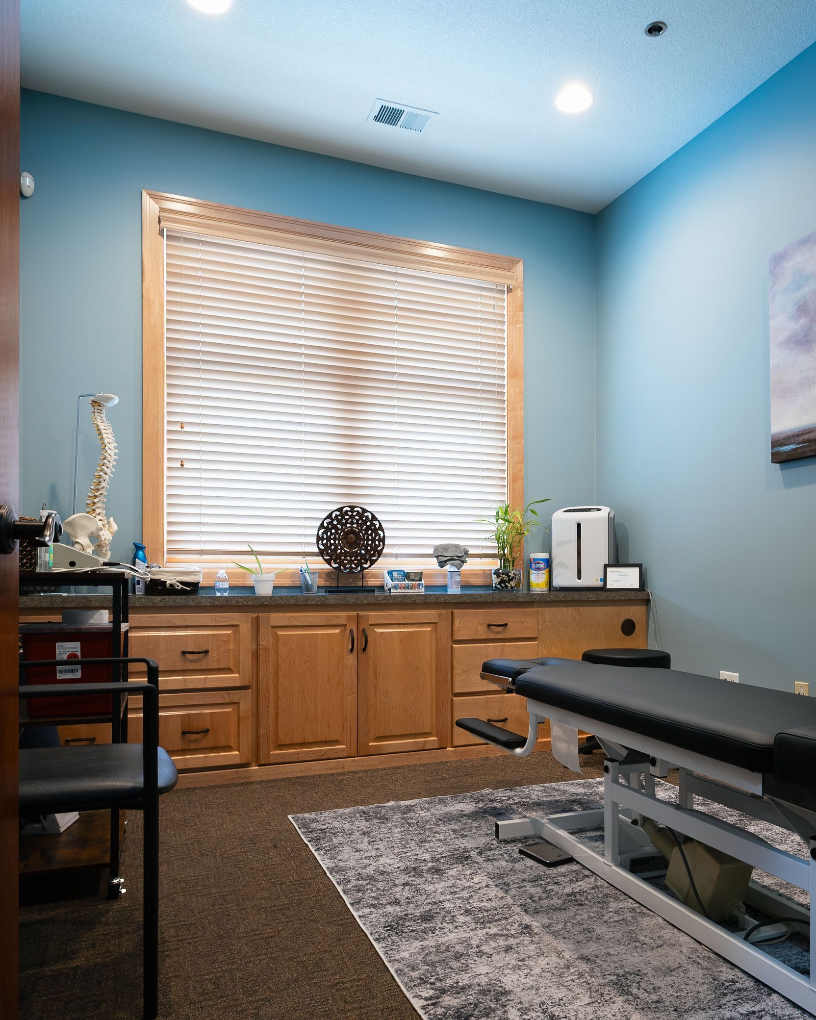 Chiropractor's office: treatment table, cabinetry, window with blinds, decorative items. Blue walls, neutral rug.