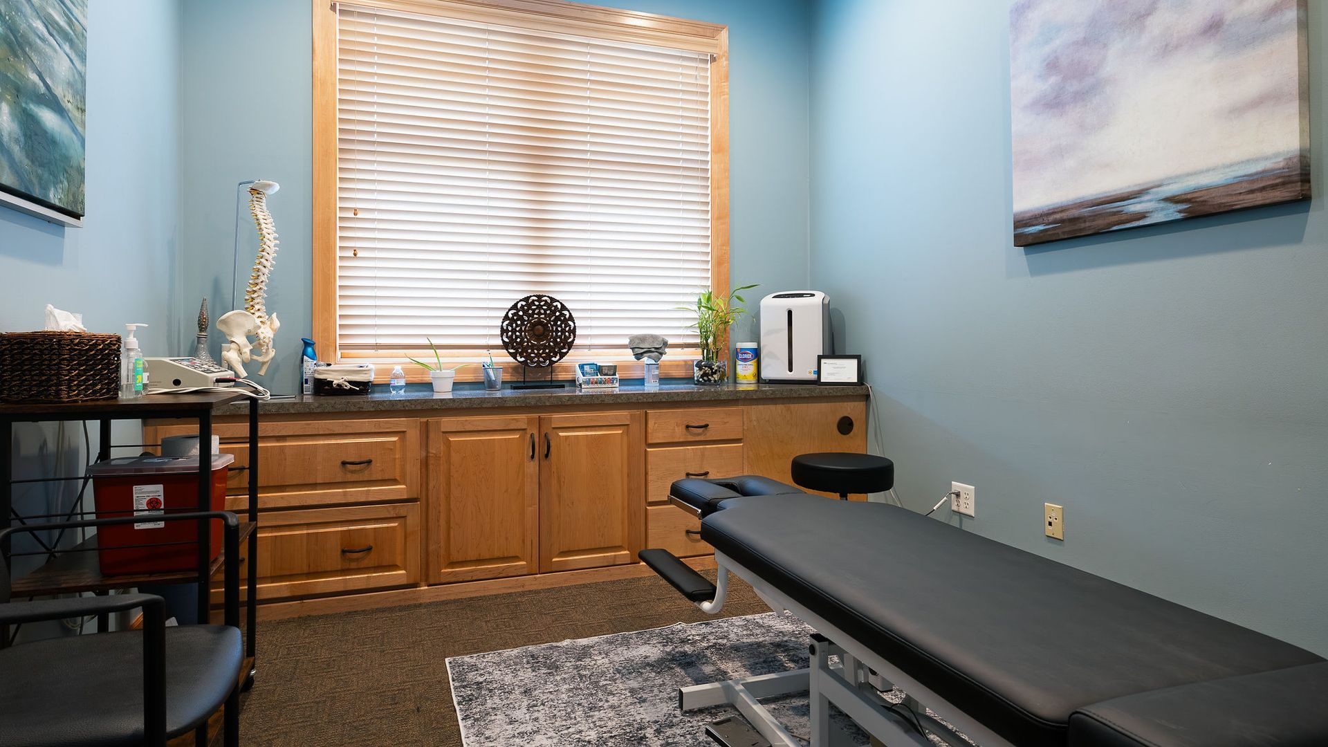 Chiropractor's office interior: treatment table, wooden cabinets, window with blinds, and decorative items.