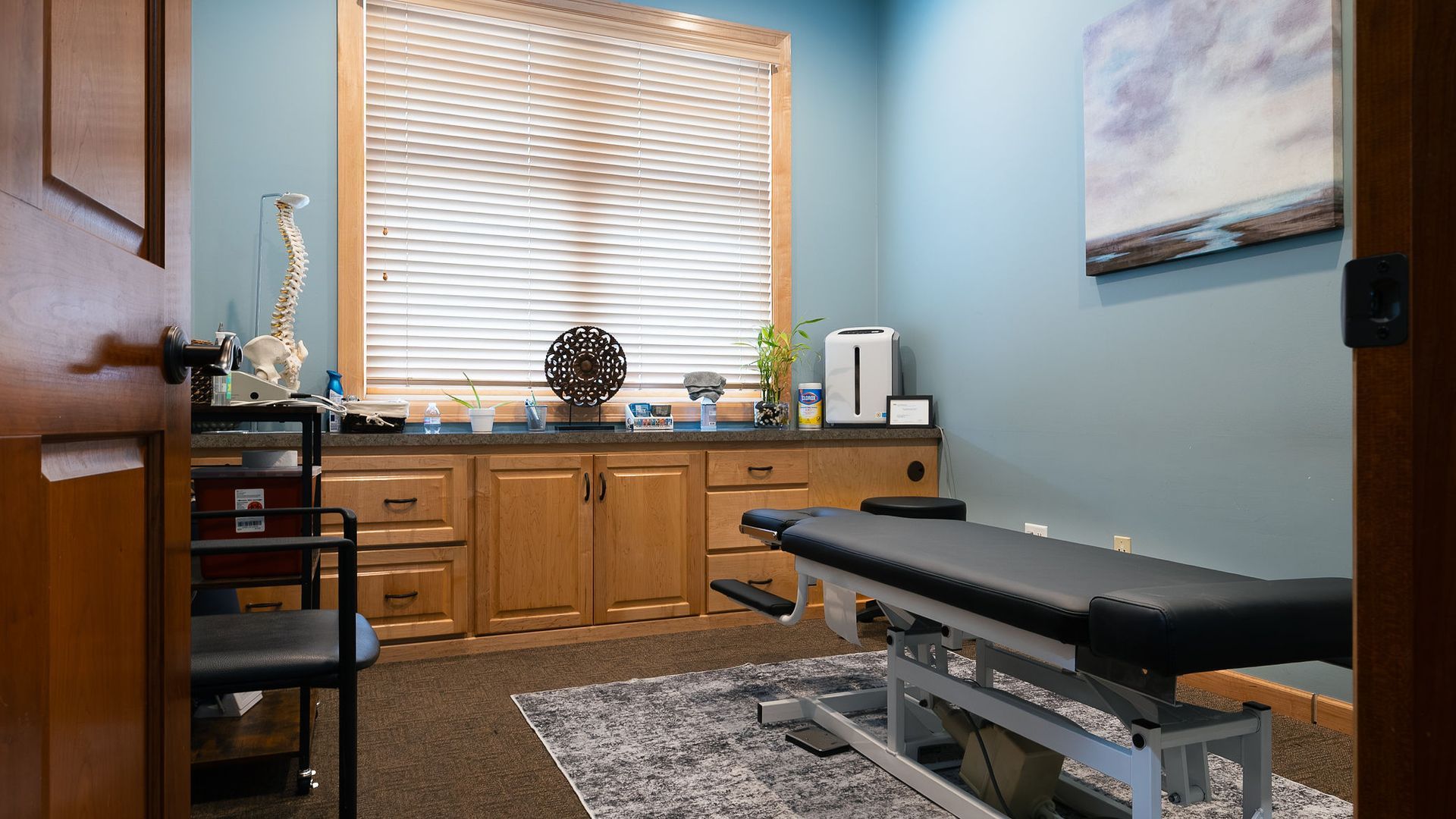 Chiropractor's office: Examination table, cabinets, window with blinds, decorations, and a door frame visible.