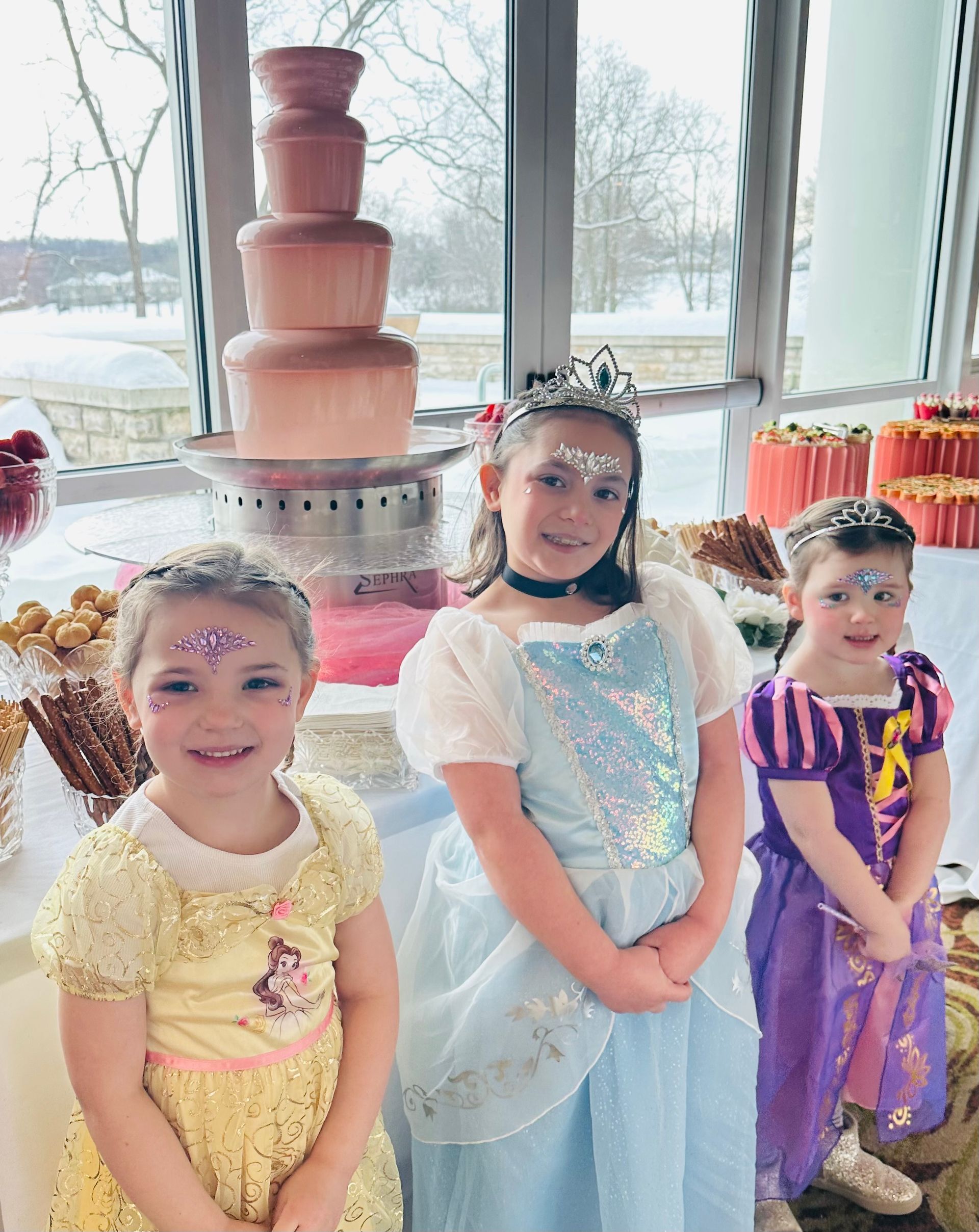 Three children in princess costumes pose smiling in front of a chocolate fountain at a party.