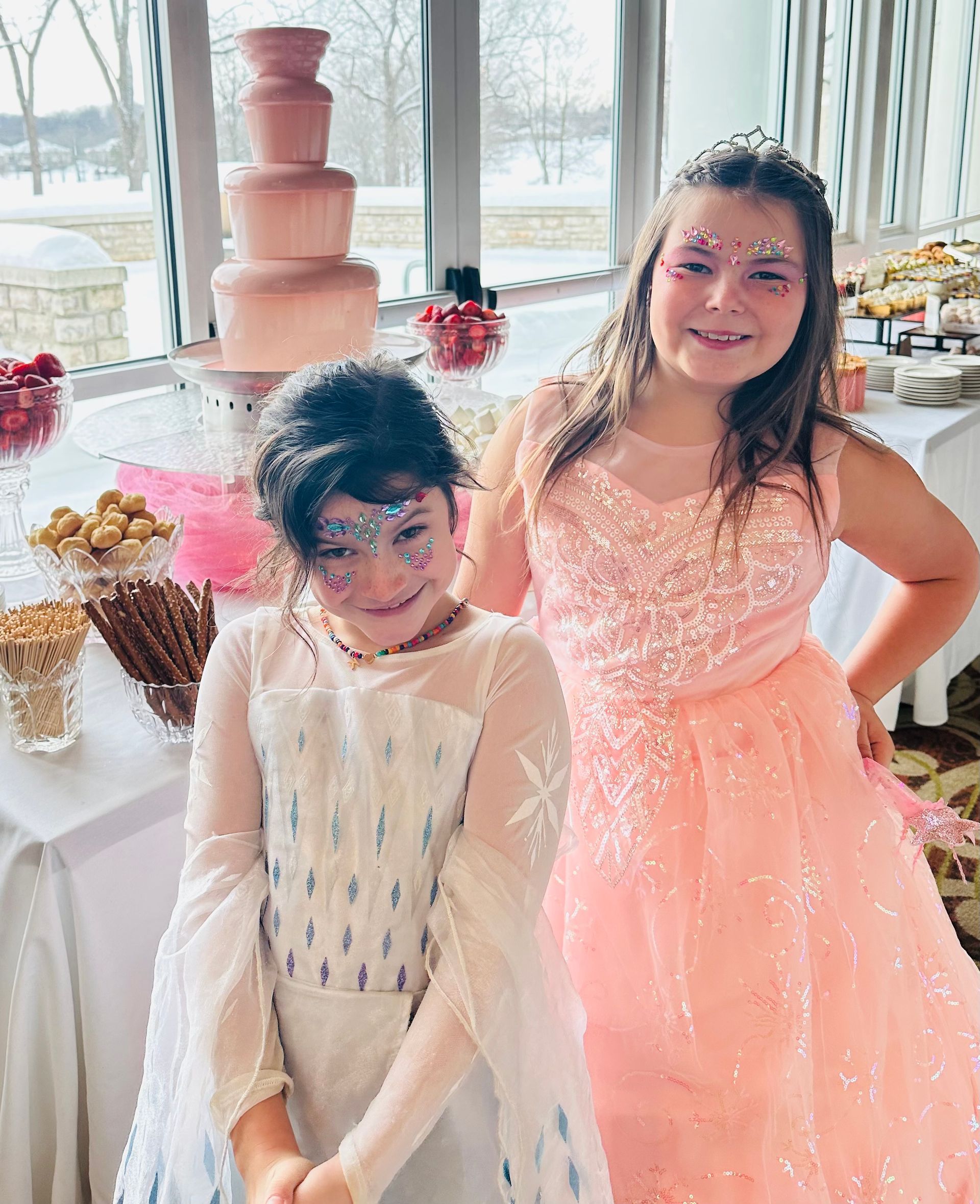 Two smiling children in formal, sparkly dresses stand in front of a party table with a pink chocolate fountain.