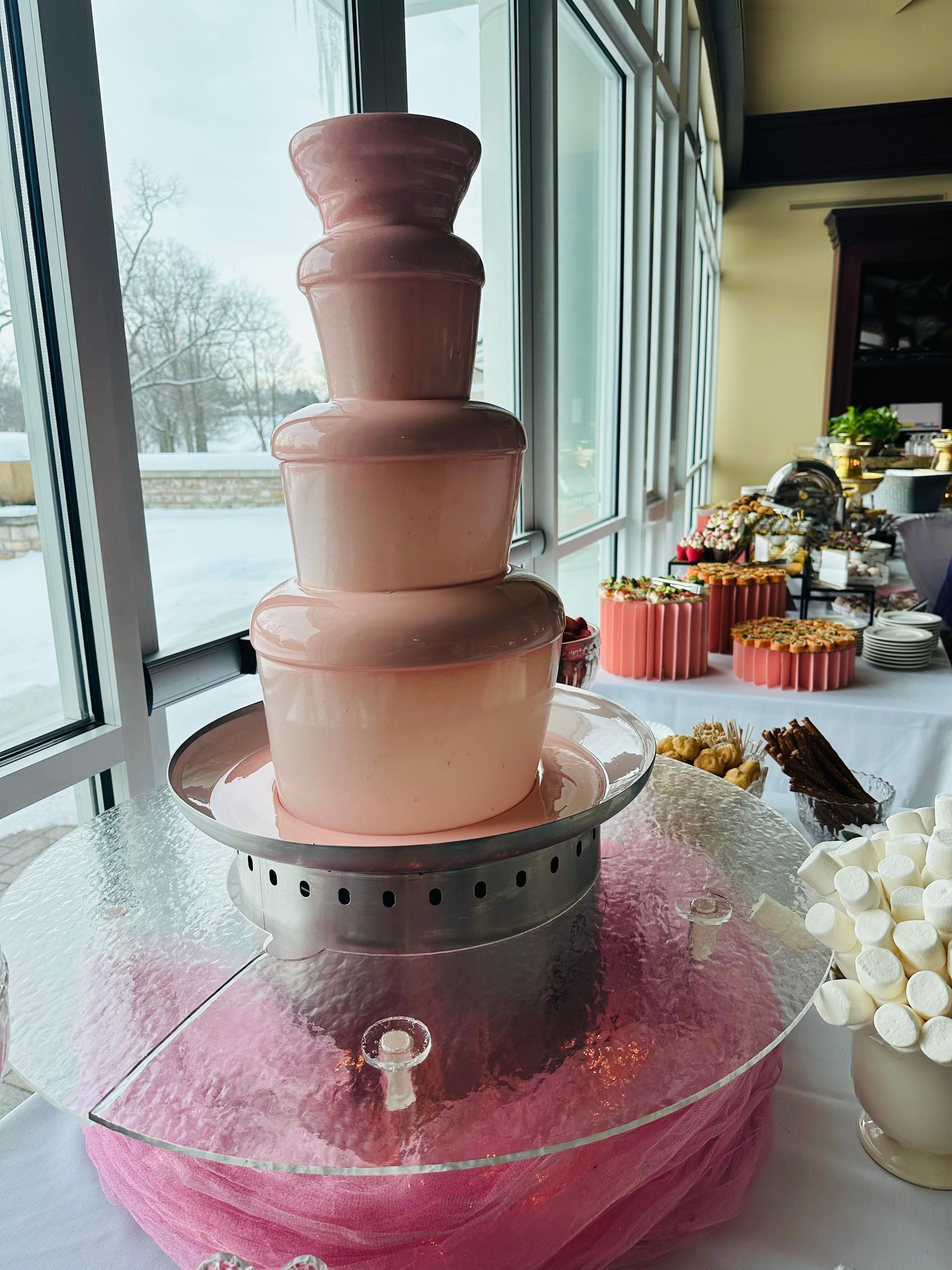 A pink chocolate fountain stands on a glass base, set on a table with snacks, with snowy trees visible through a window.