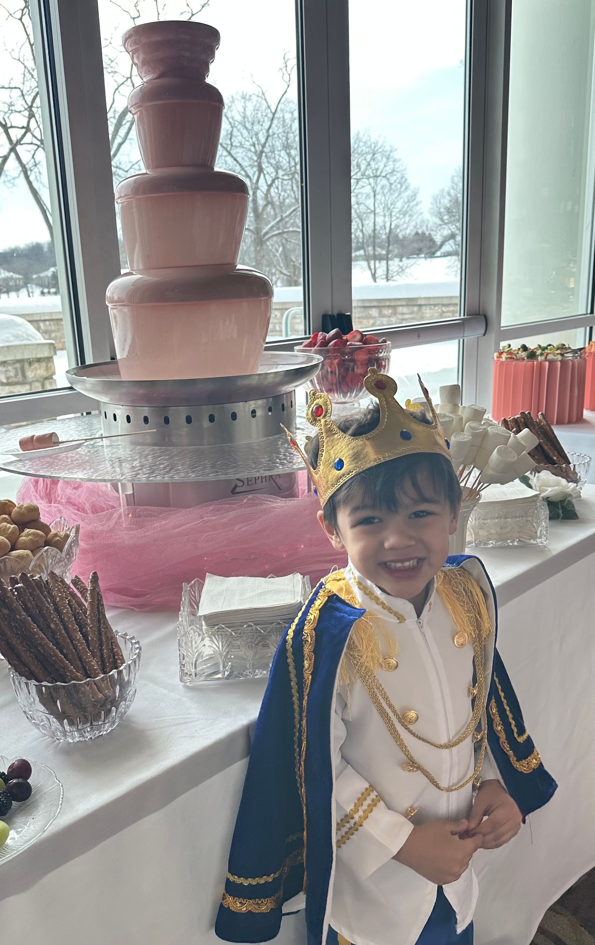 A smiling child in a royal costume and crown stands at a table featuring a tall pink chocolate fountain and snacks.