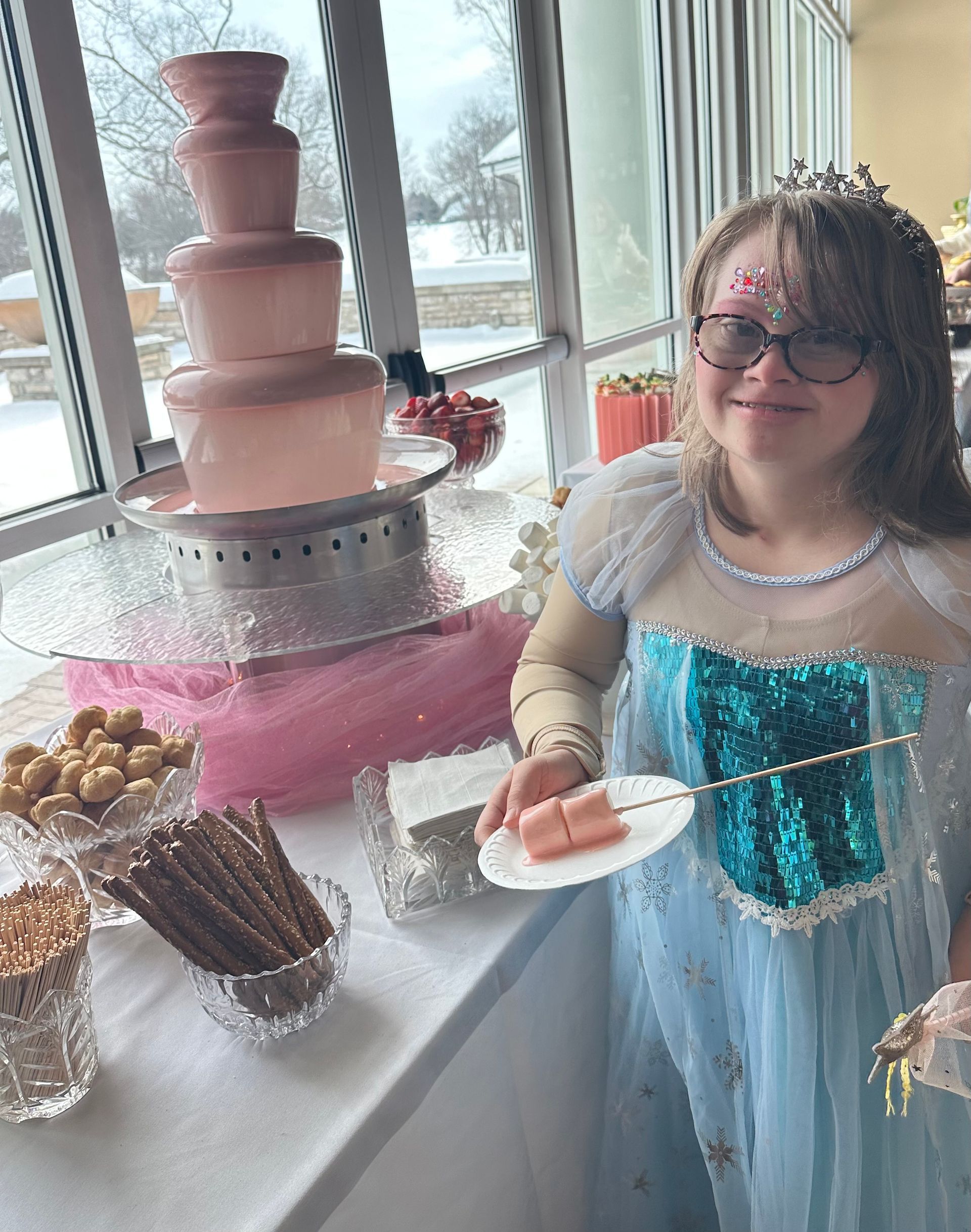 A person in a blue costume stands smiling next to a pink chocolate fountain and a table of treats.