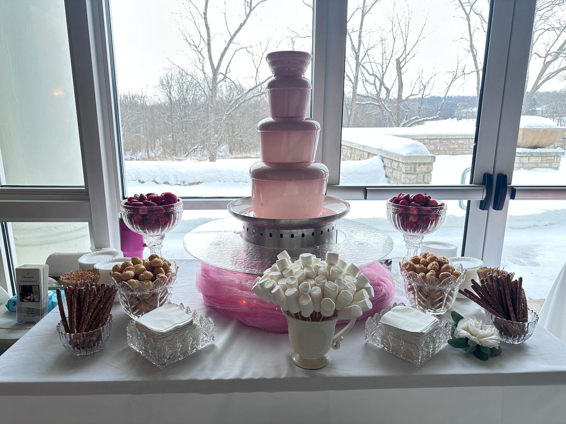 A pink chocolate fountain on a white table surrounded by crystal bowls of pretzels, marshmallows, and berries.