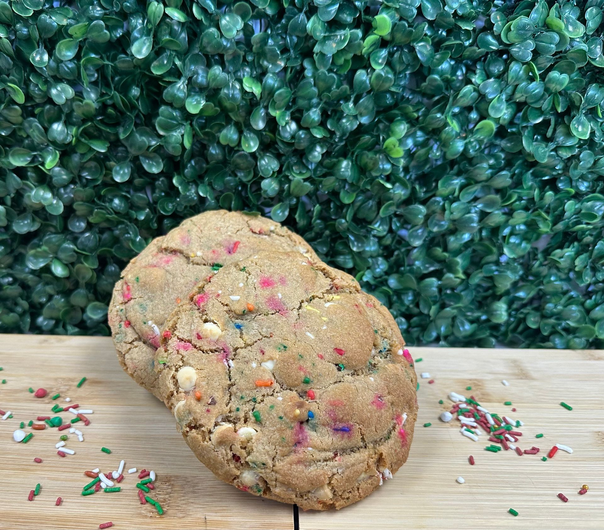 A funfetti cookie with white chocolate chips on a wooden surface with a green leafy wall background.