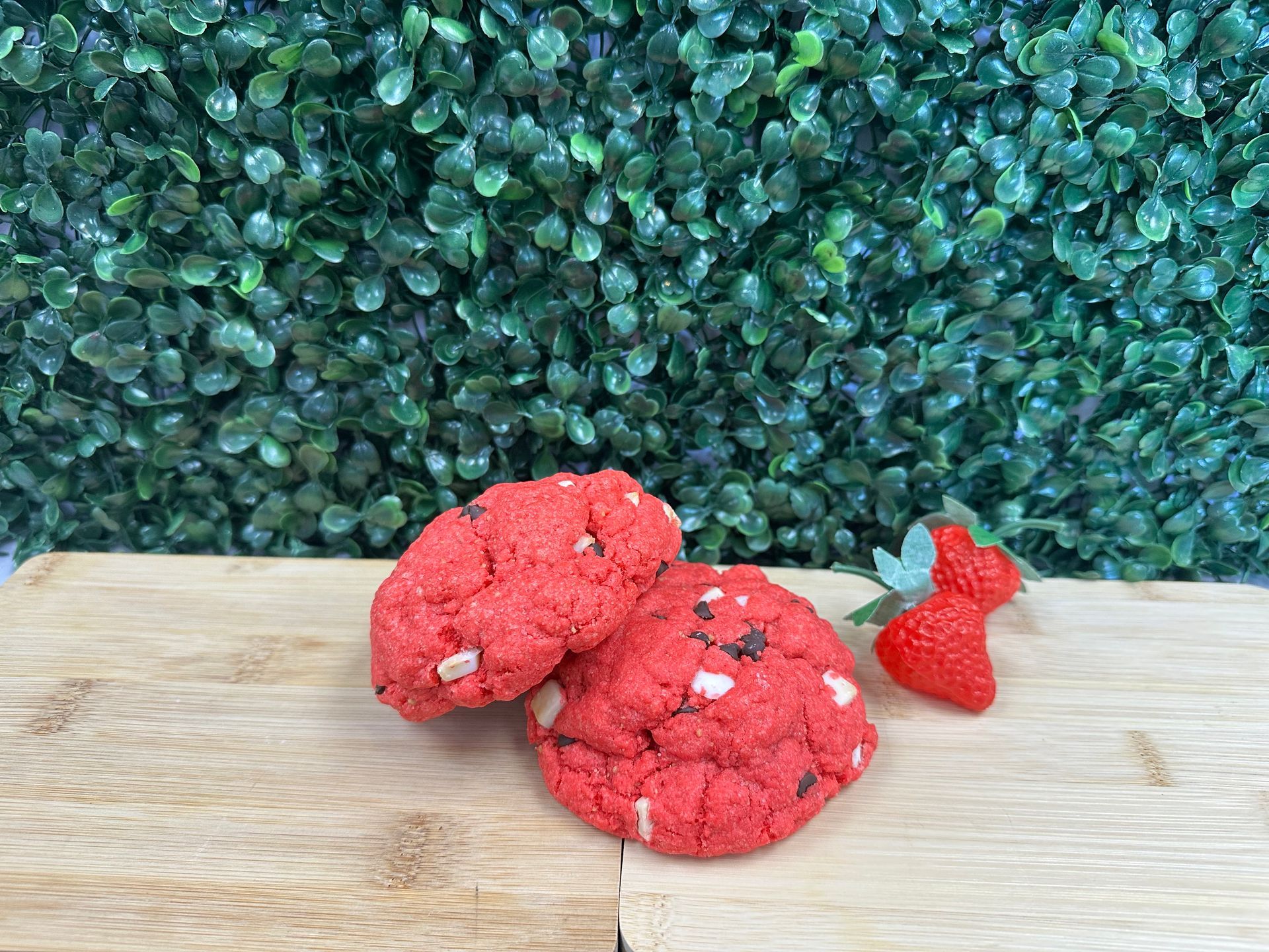 Two red cookies with white chocolate chips sit on a wooden surface next to a single fresh strawberry against green plants.