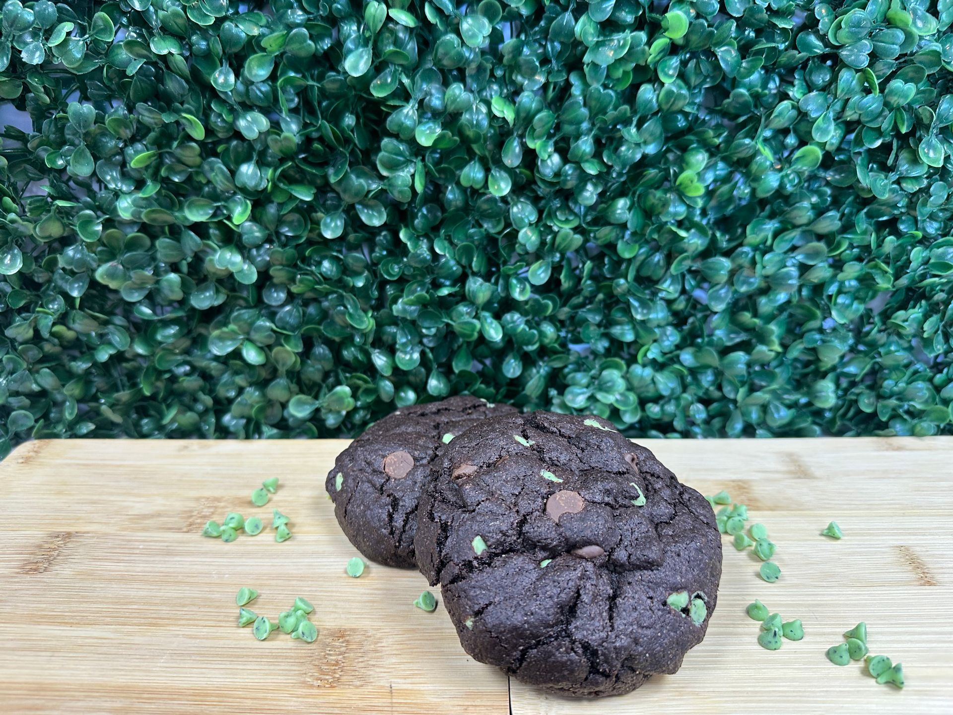 Two dark chocolate mint cookies with green sprinkles sit on a wooden cutting board against a lush green leafy background.