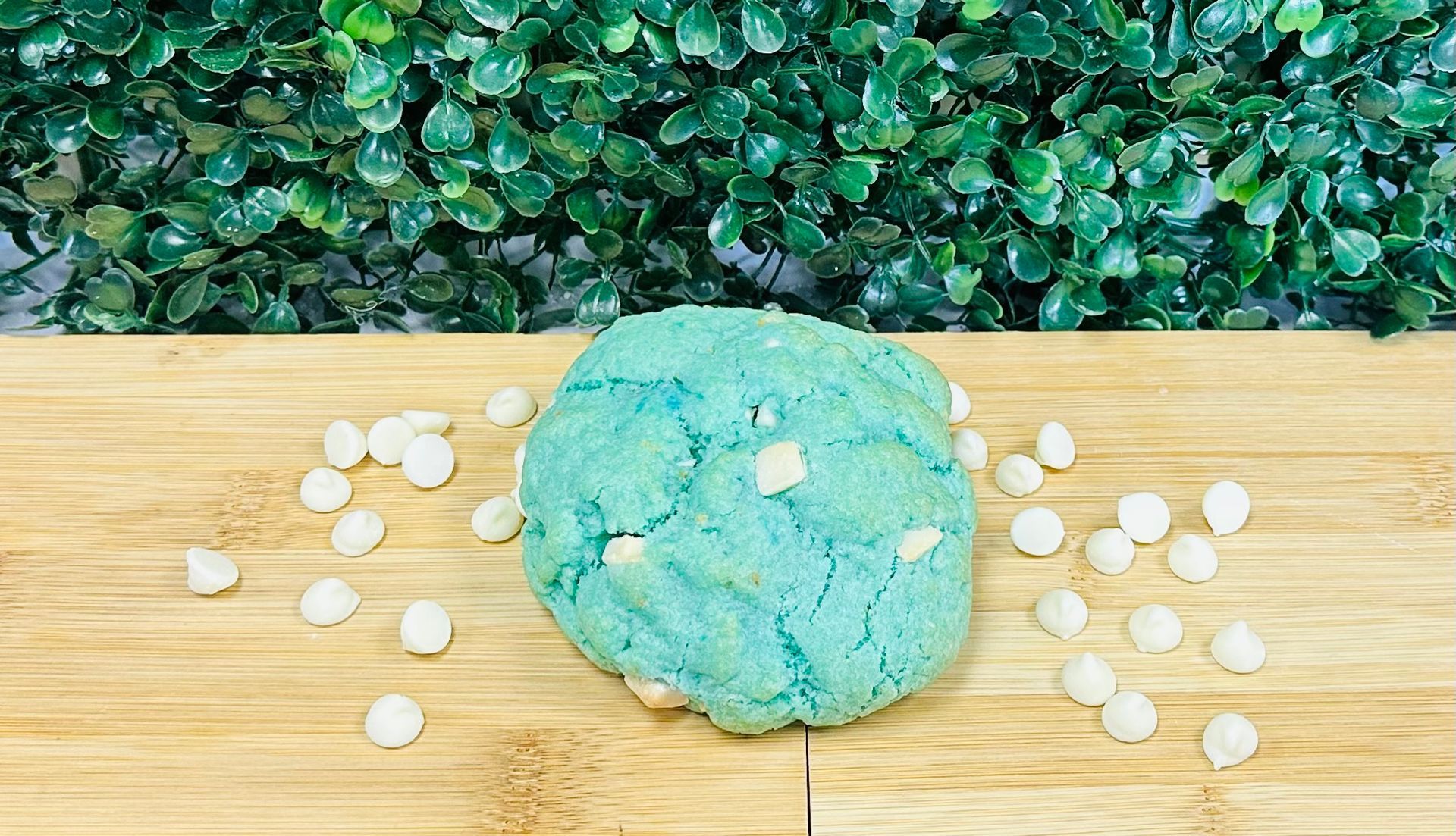 A bright blue cookie with white chocolate chips sits on a wooden cutting board in front of a green leafy hedge.