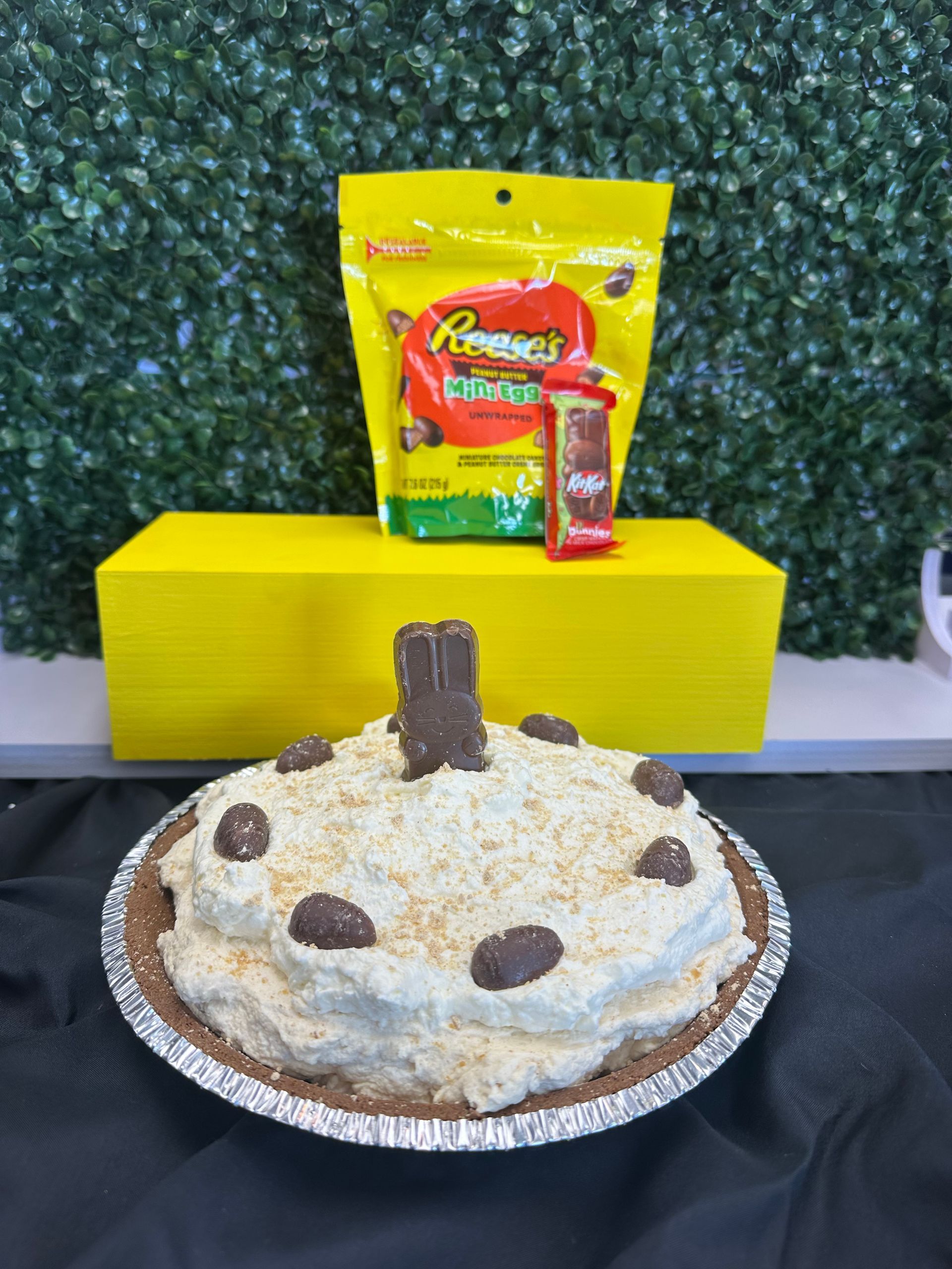 A creamy pie topped with chocolate candy, displayed on a black surface in front of a yellow box and a bag of Reese's.