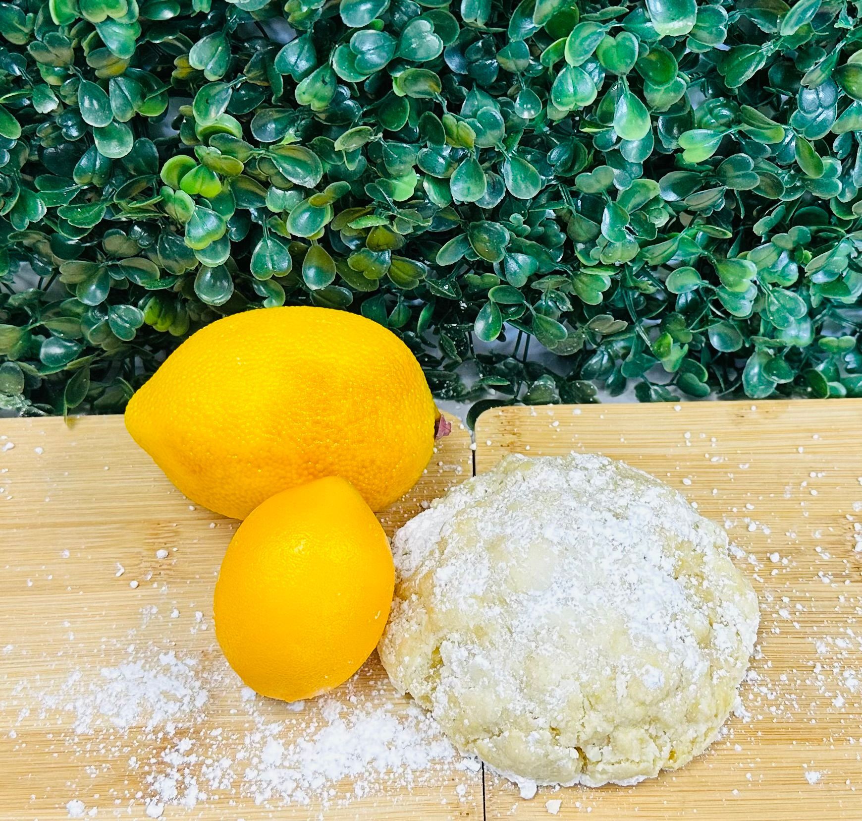 Two lemons and a powdered cookie sit on a light wood board in front of a green leafy background.